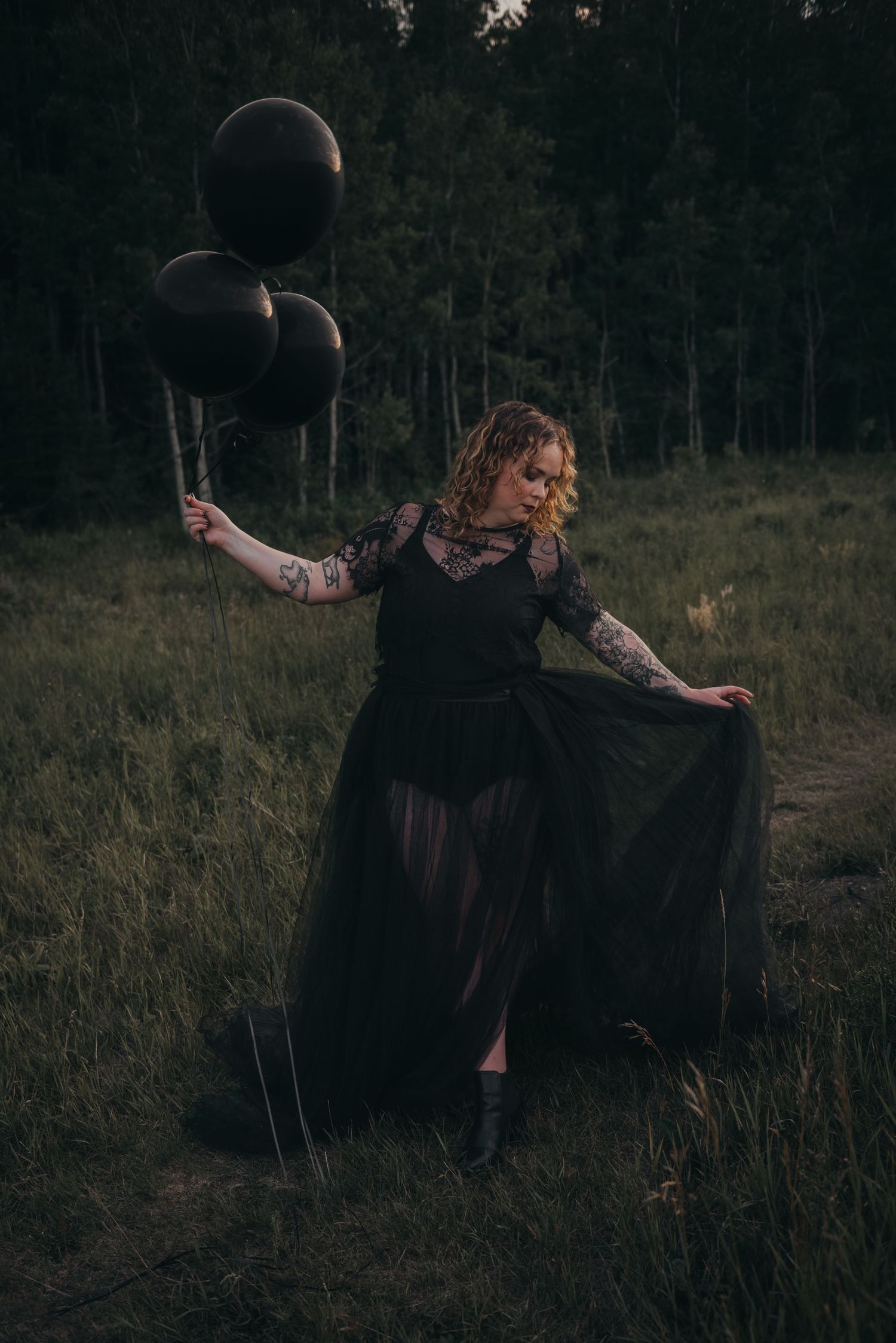 Woman having photoshoot in black lace gown holding black balloons in a dark Calgary forest meadow at dusk.