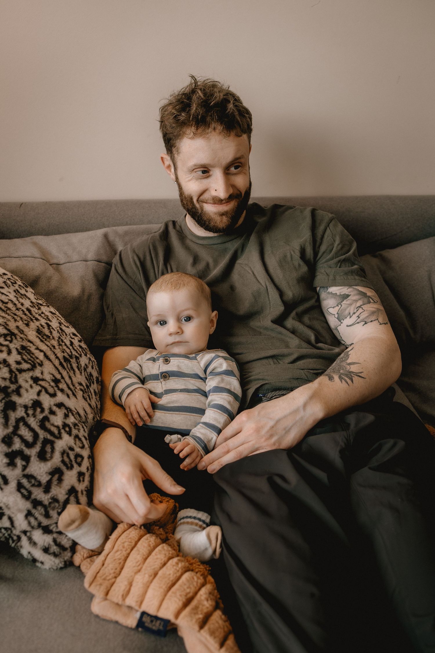Smiling bearded father with tattoos during a family photo session sitting on gray sofa holding his baby in striped onesie at home.