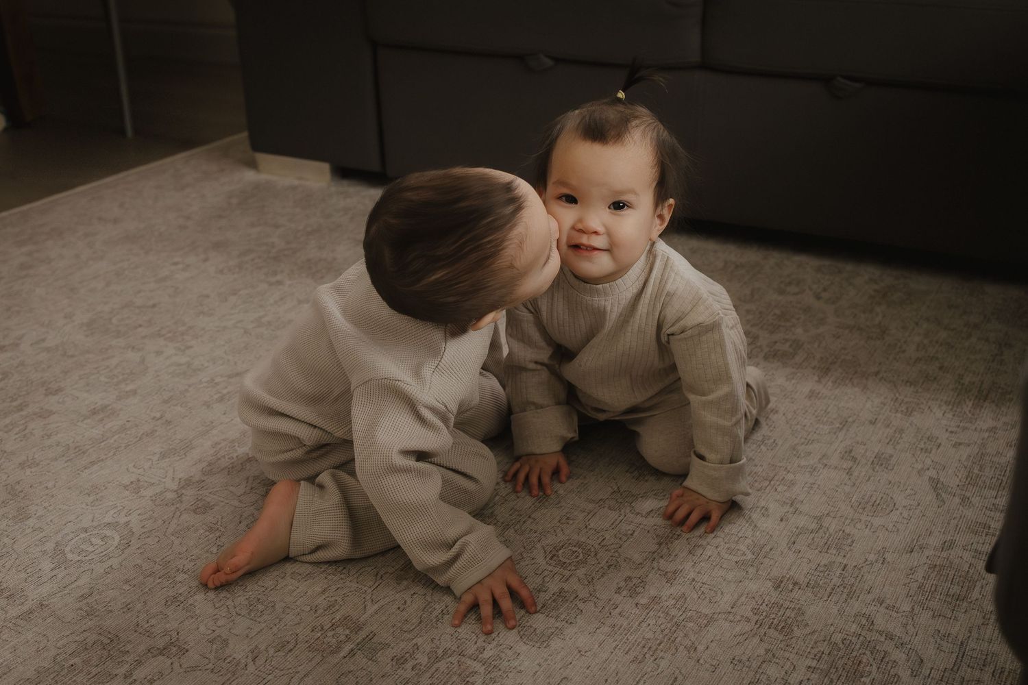 Two young siblings in matching beige outfits sitting on a carpet, older child gently kissing baby on the cheek.