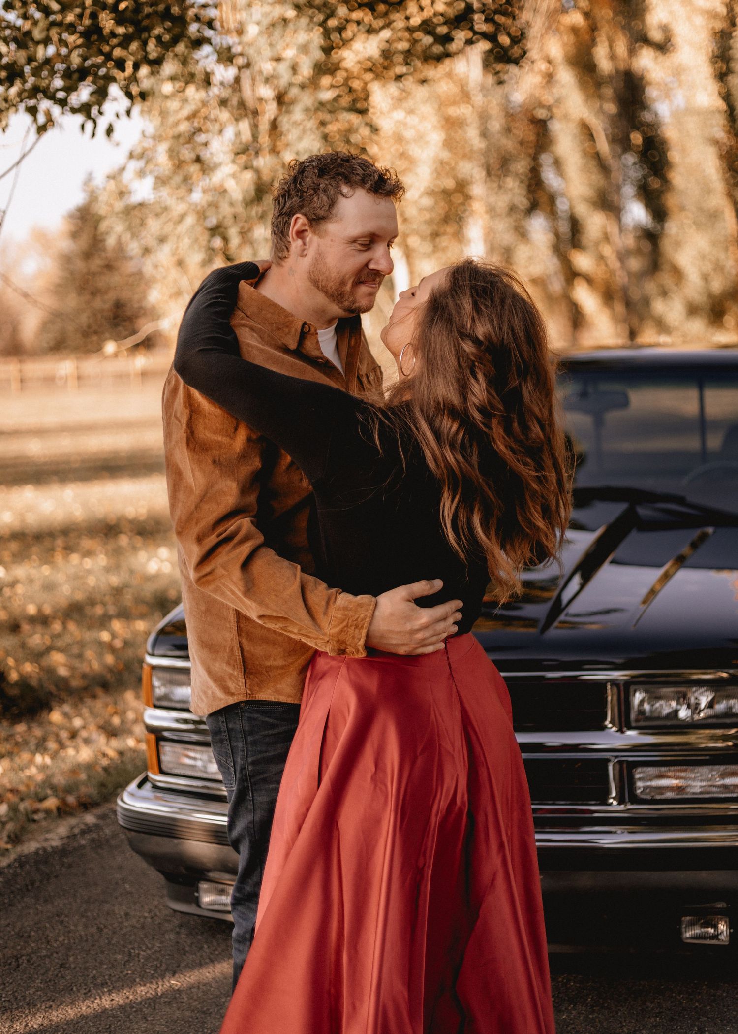 High River couple embracing near a vintage black truck surrounded by autumn trees, woman in red skirt and man in brown jacket.