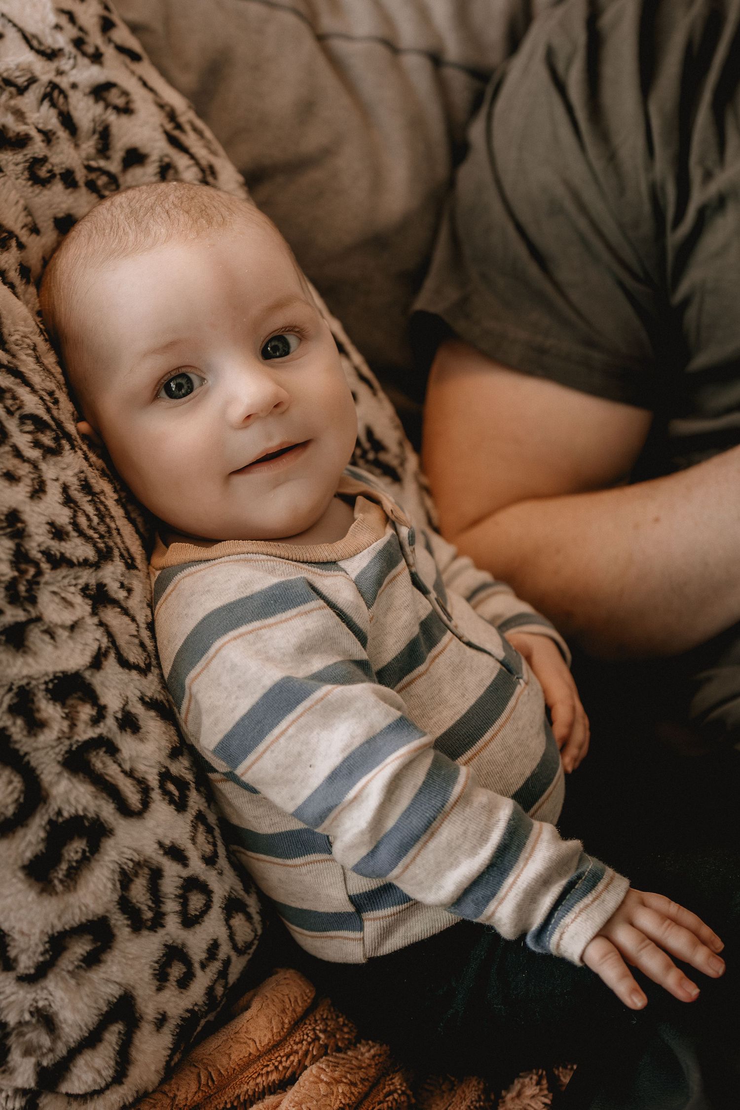 Adorable baby in striped onesie sitting on a couch with leopard print pillow, looking up with curious blue eyes at family photographer