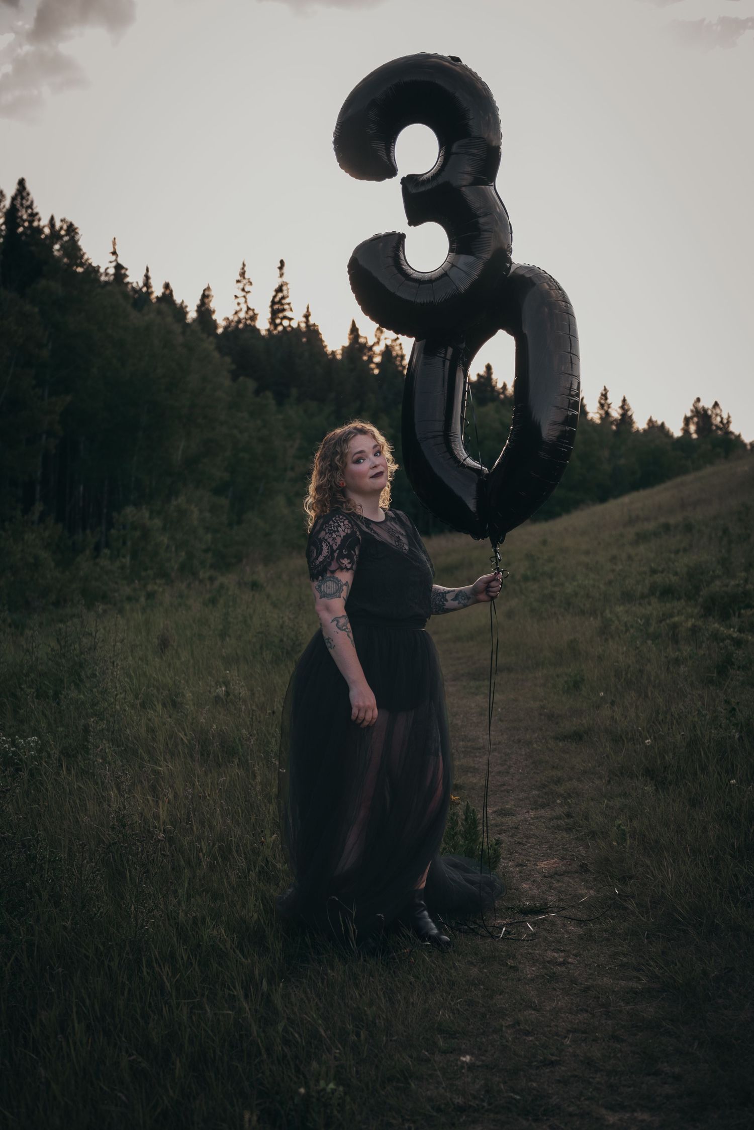 Woman in black dress holding large black number 30 balloons outdoors at dusk, celebrating 30th birthday.