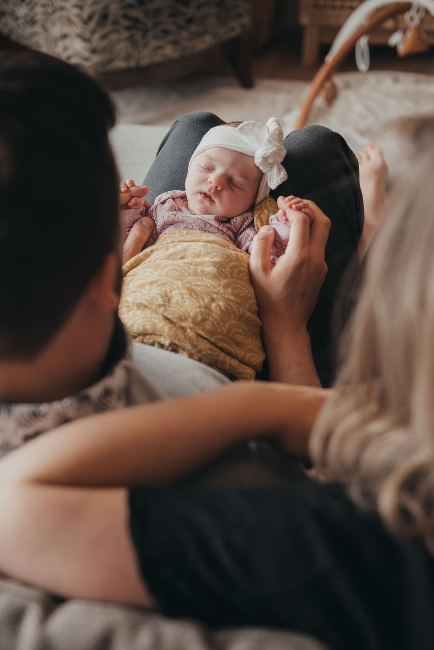 Parents gazing lovingly at their sleeping newborn baby wearing a white bow headband, wrapped in a yellow blanket.