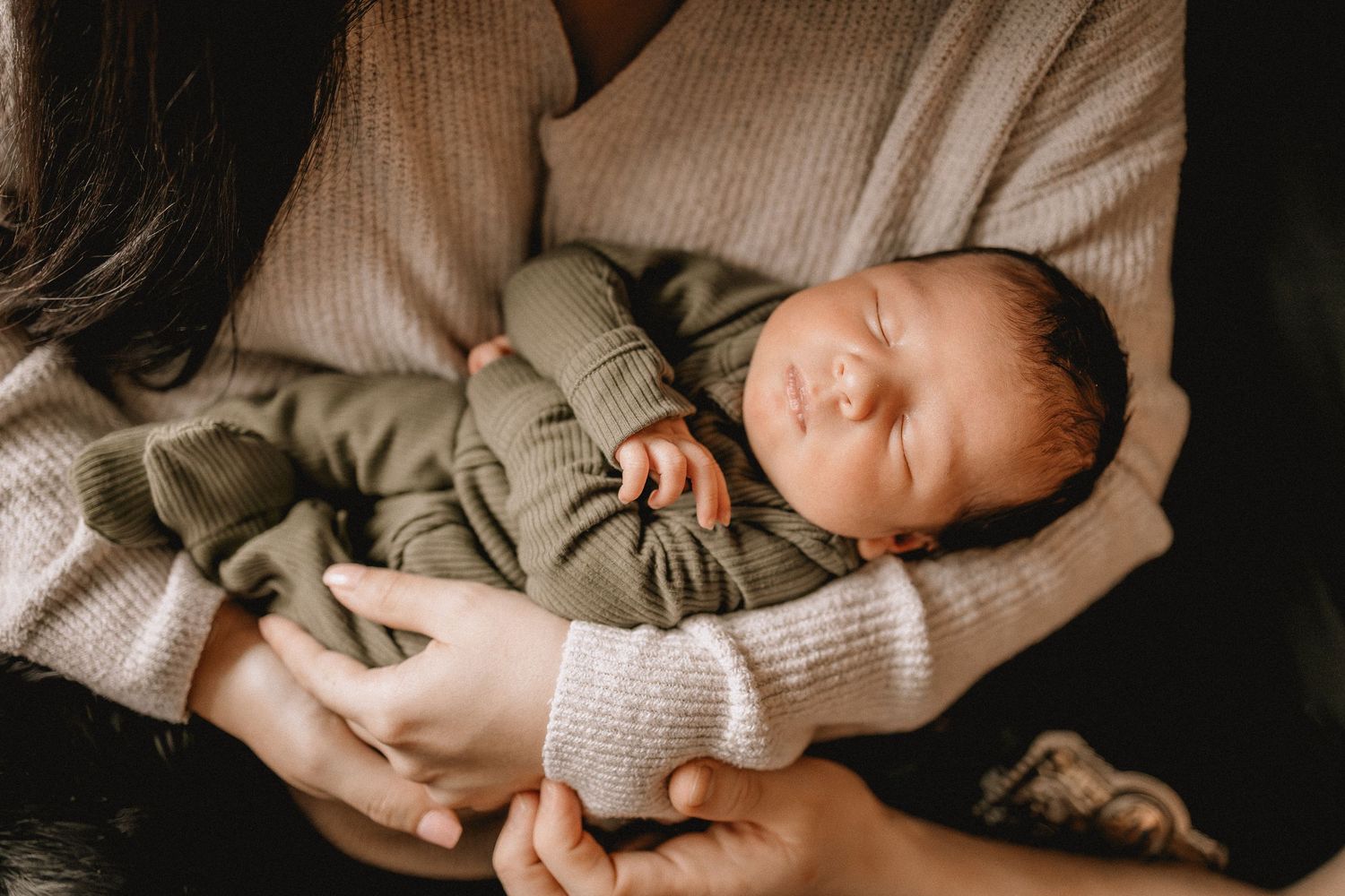 A sleeping newborn baby wearing a sage green outfit rests peacefully in someone's arms wearing a cream sweater.