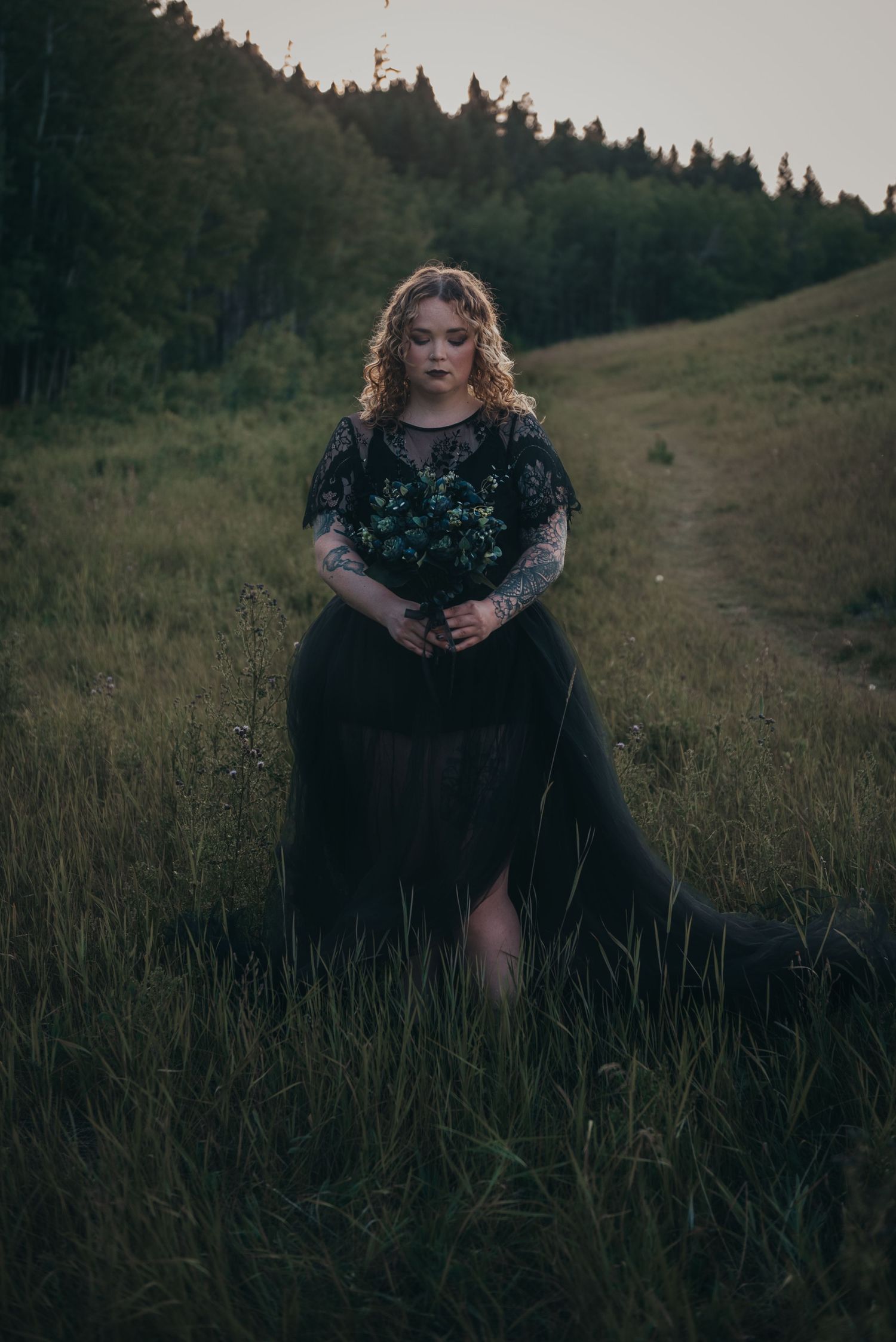 Woman in black gothic gown holding dark bouquet standing in lush green meadow in Shannon Terrace at dusk.