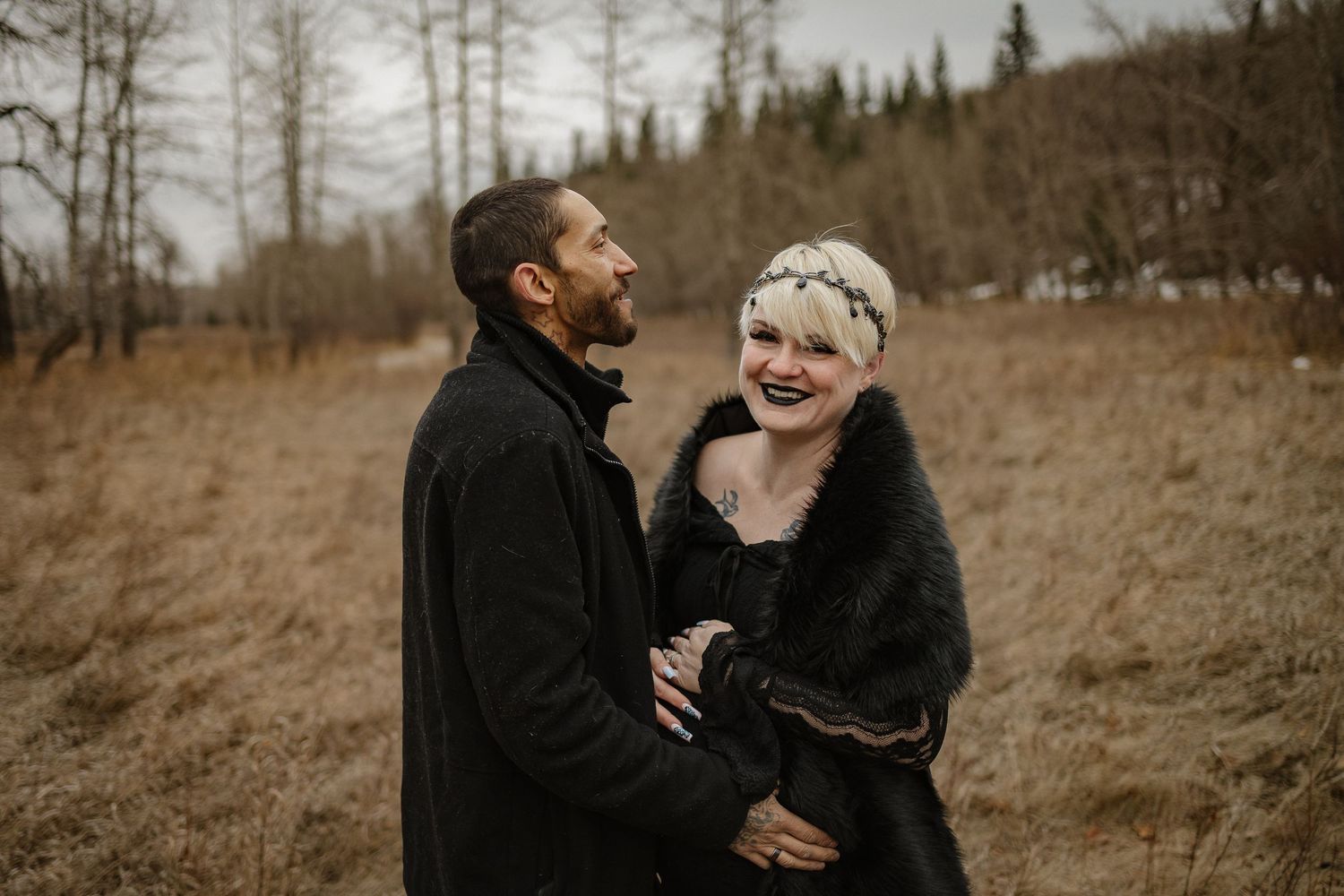 Couple laughing together outdoors in autumn field, woman wearing floral headpiece and black fur wrap, man in black coat.
