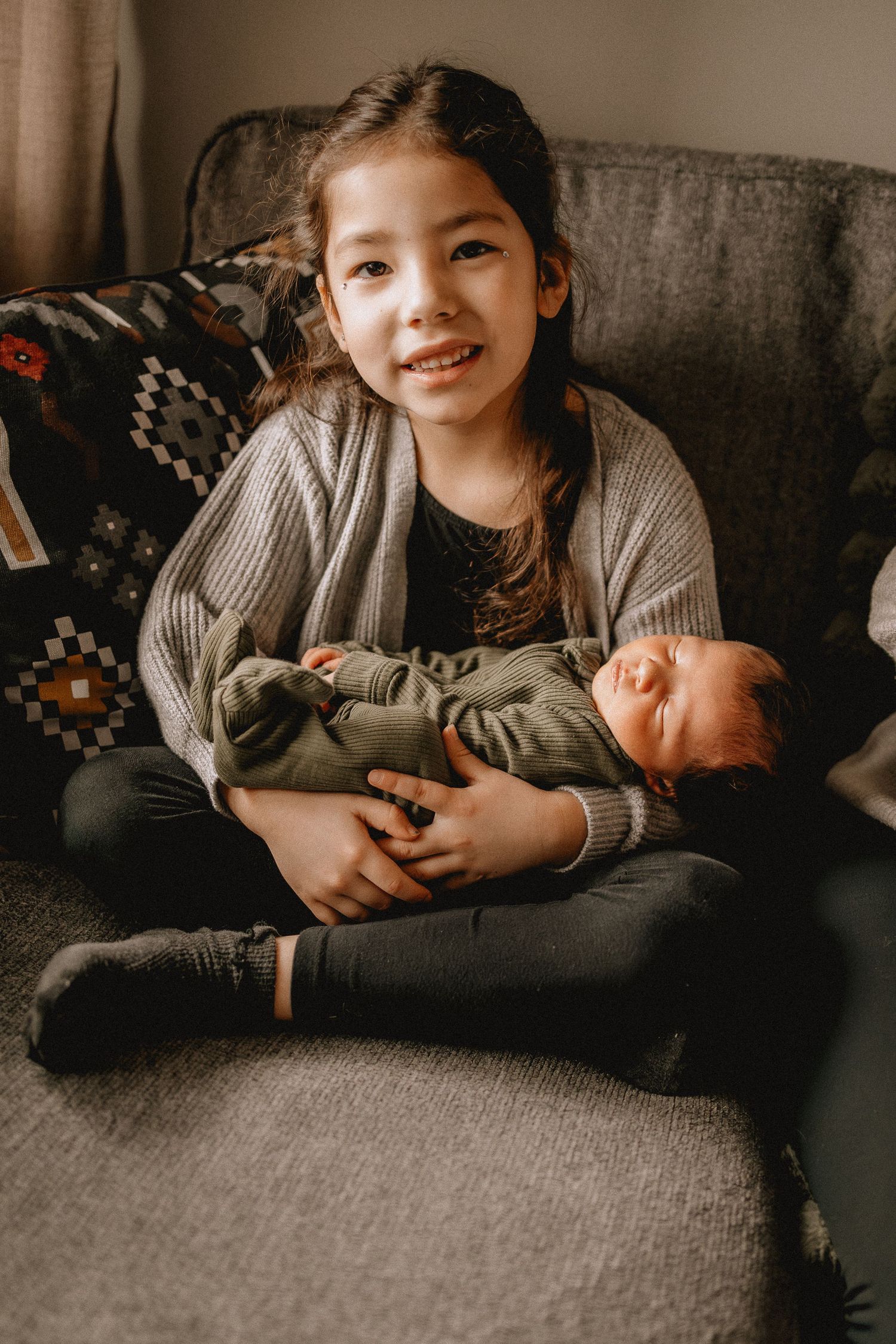 Young girl sits cross-legged on gray sofa, smiling while holding sleeping baby dressed in green outfit for newborn photoshoot