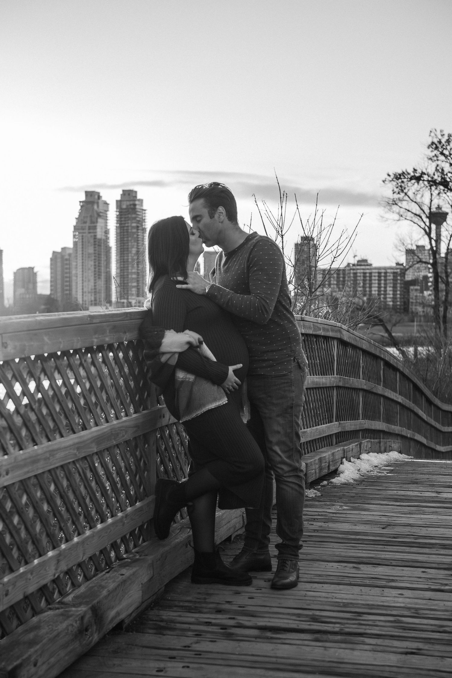 Couple sharing intimate moments on a wooden bridge with city skyline in black and white maternity photography.