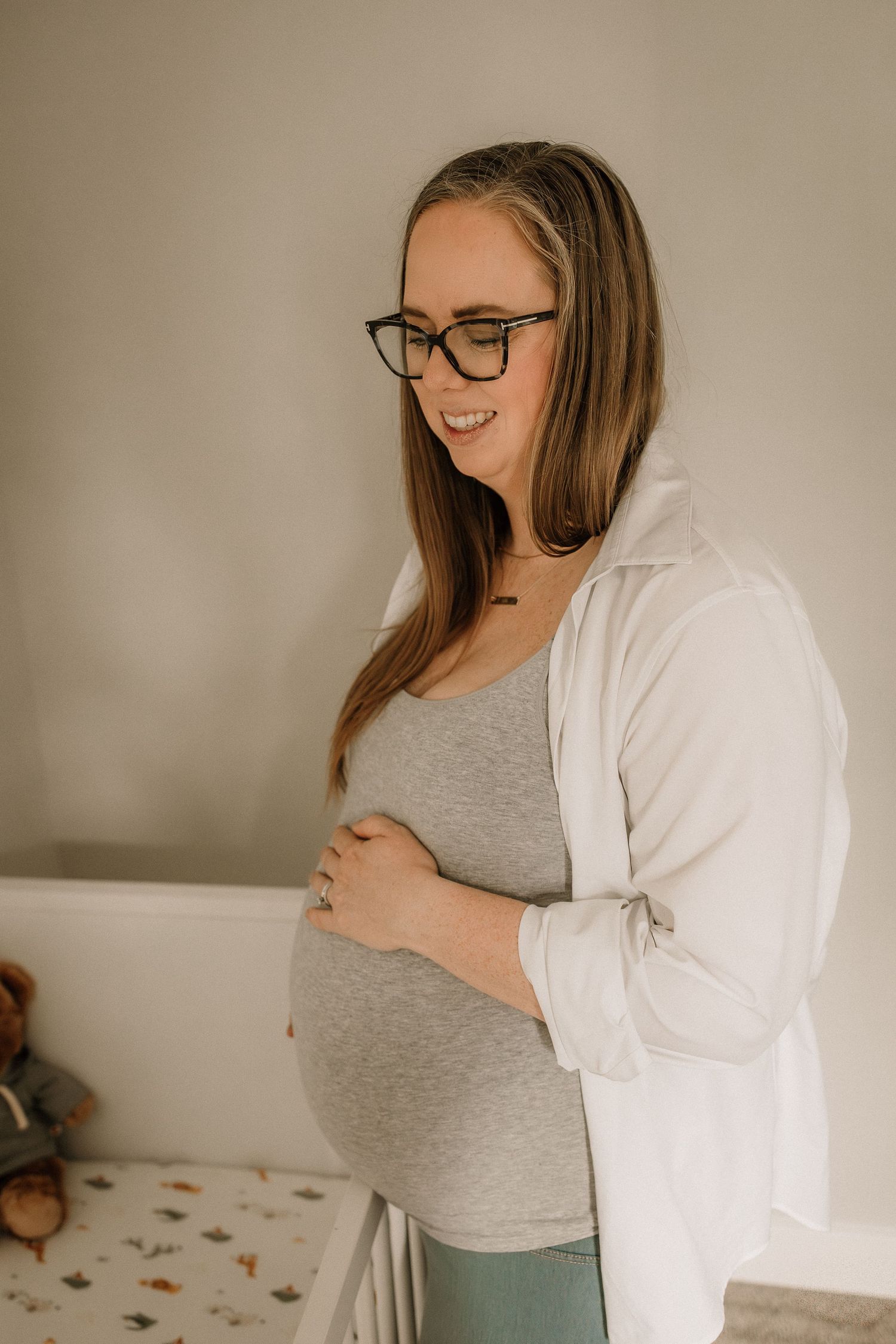 Smiling pregnant woman in glasses and gray top cradling her baby bump in a cozy nursery room during a maternity photography session.