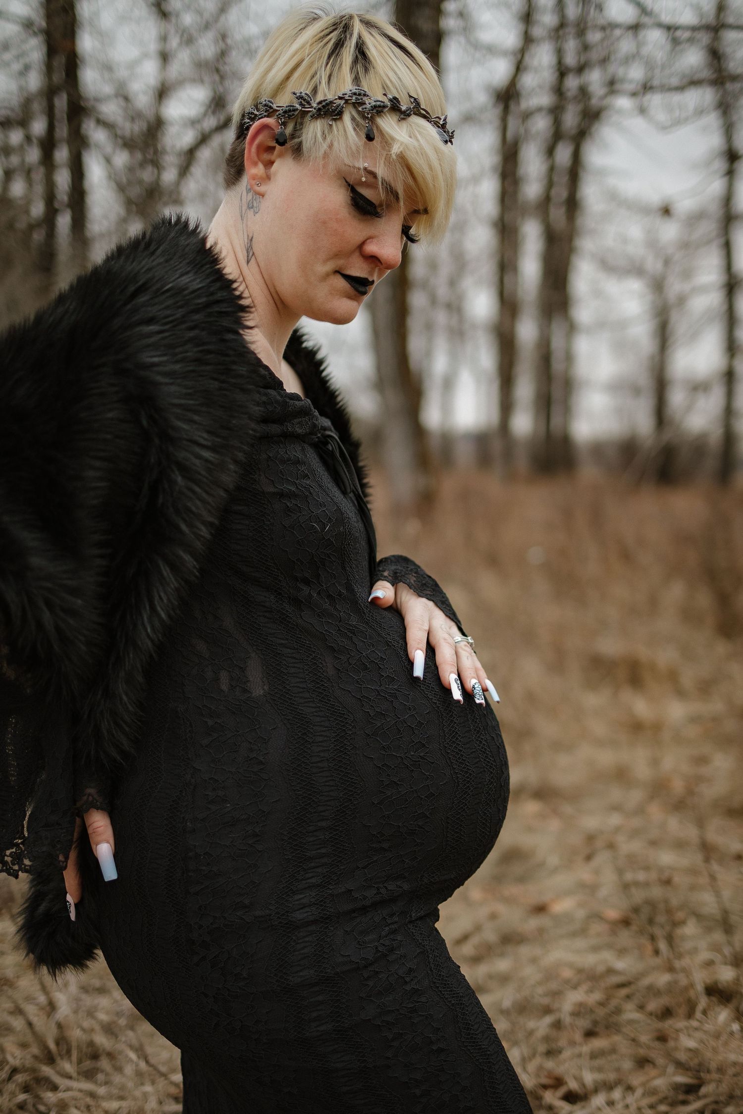 Pregnant woman in black dress and fur wrap with floral crown poses outdoors in a moody forest setting.