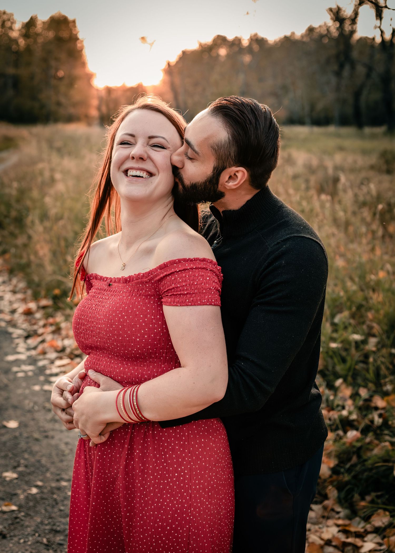 A couple shares a romantic sunset moment outdoors, man in black kissing womans cheek as she laughs in red dress.