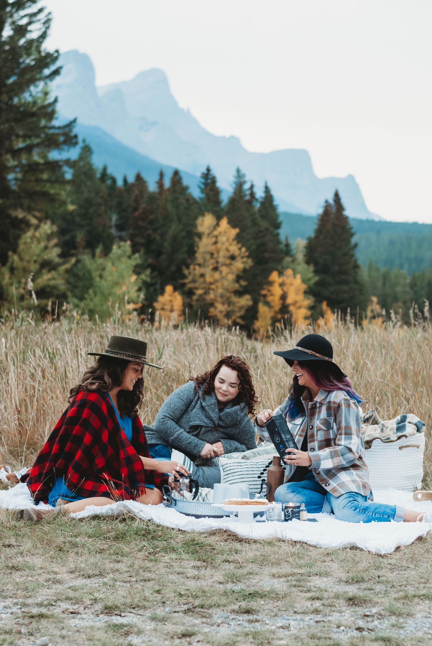 Three women in flannel and hats enjoy an autumn picnic on a blanket in a Canmore mountain meadow with evergreens and peaks behind.
