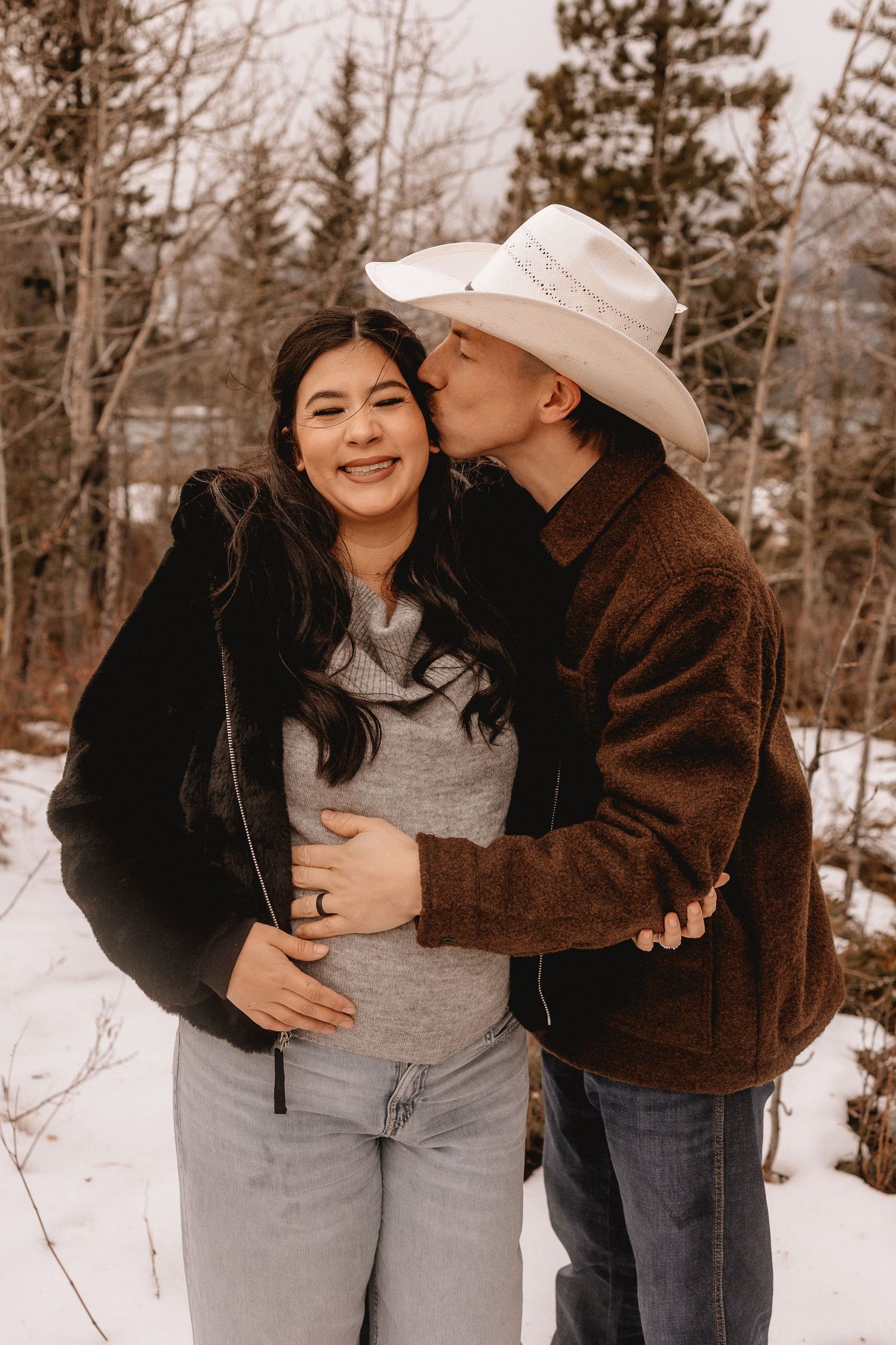 A couple shares tender moments in a snowy winter forest setting wearing casual outerwear and jeans.