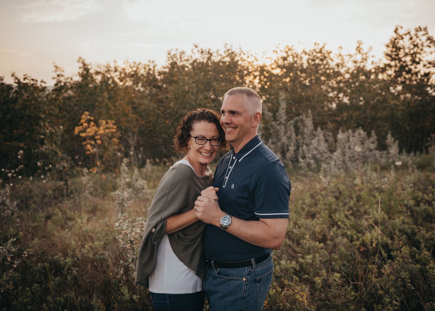 A smiling couple embraces outdoors in a golden Calgary field with autumn foliage, wearing casual attire in warm evening light.