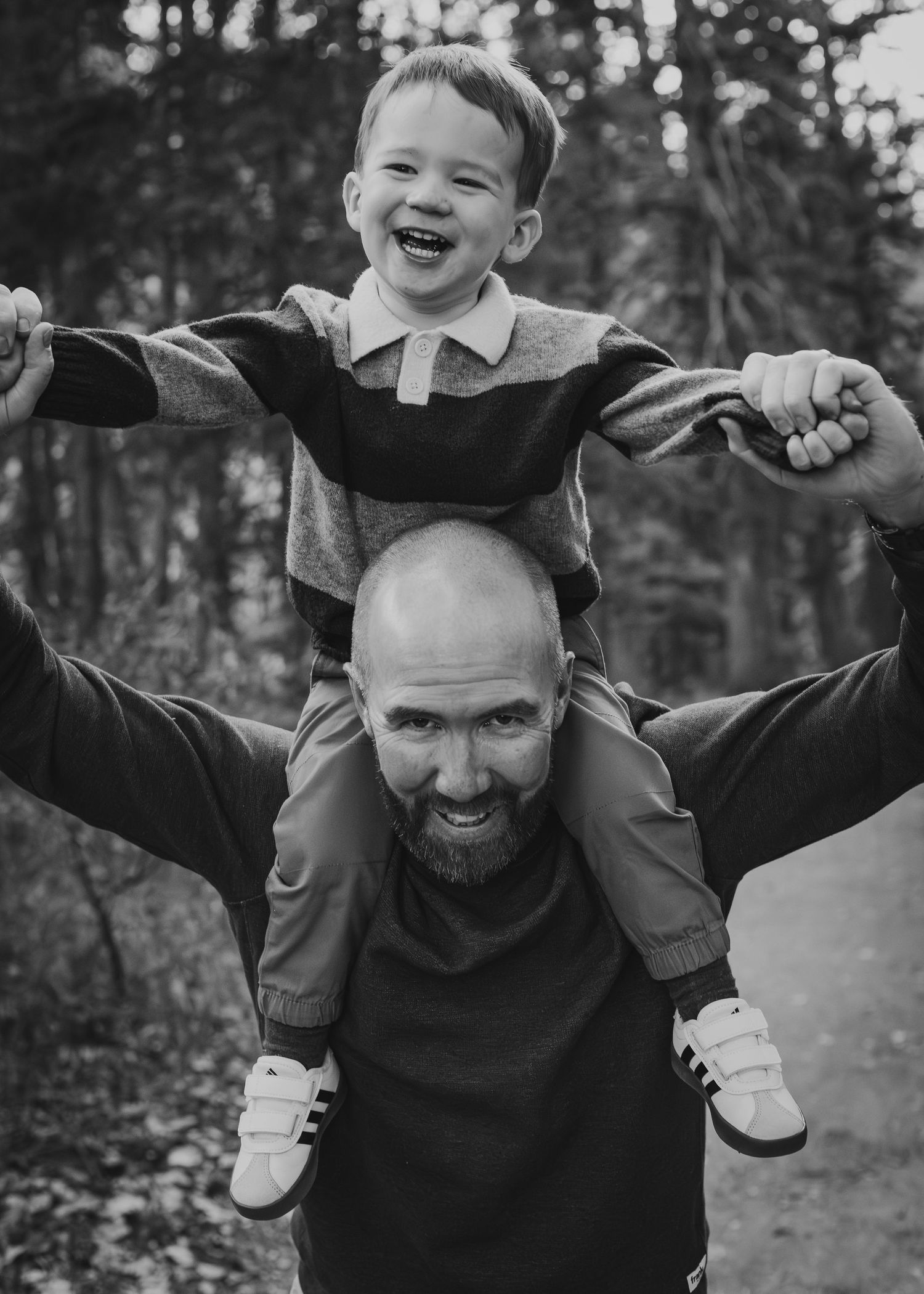 Bald bearded man smiling with laughing young boy sitting on his shoulders in outdoor Calgary in black and white photo.
