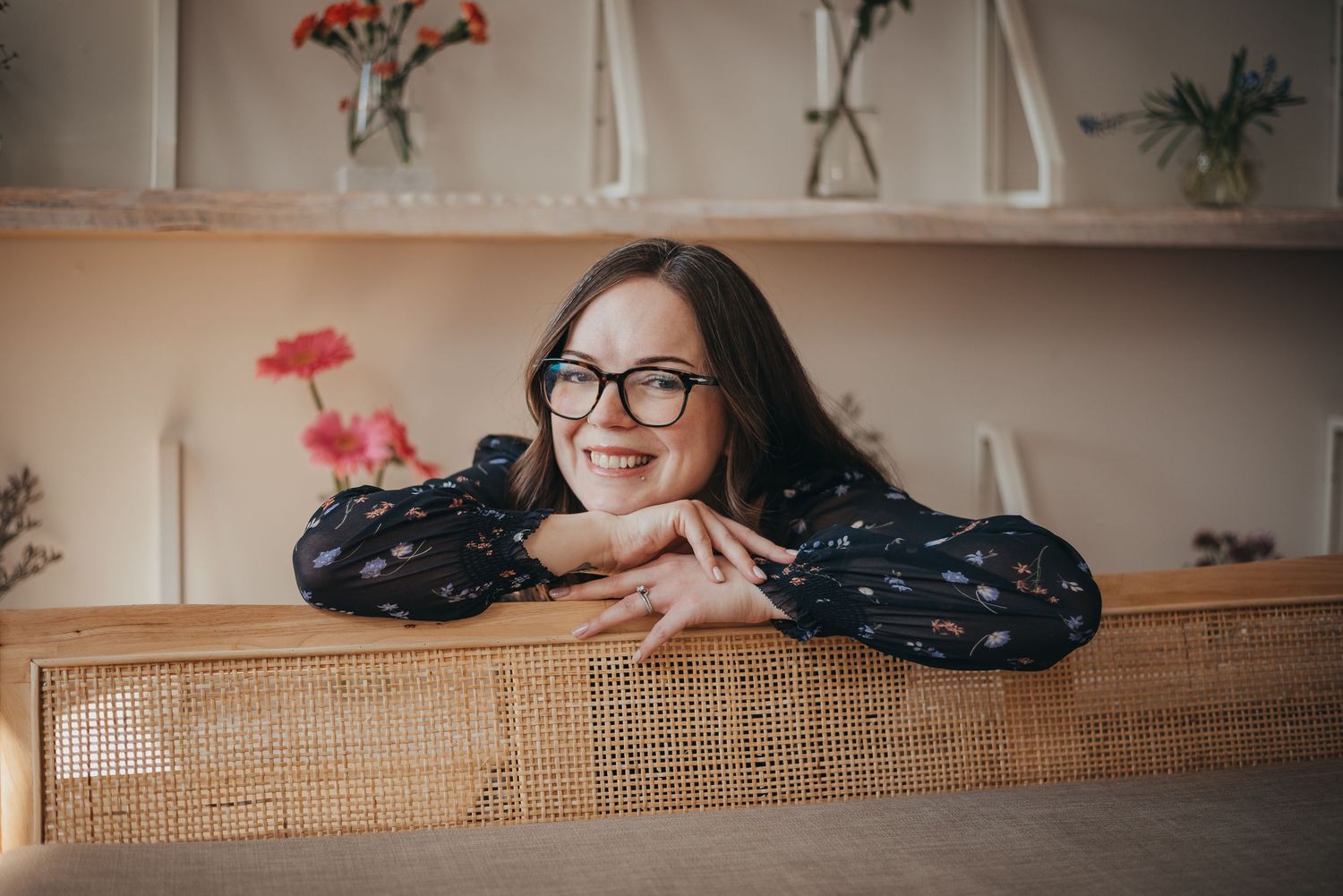 Woman with glasses smiling and leaning on a wicker couch in a cozy Calgary studio with flowers in the background.