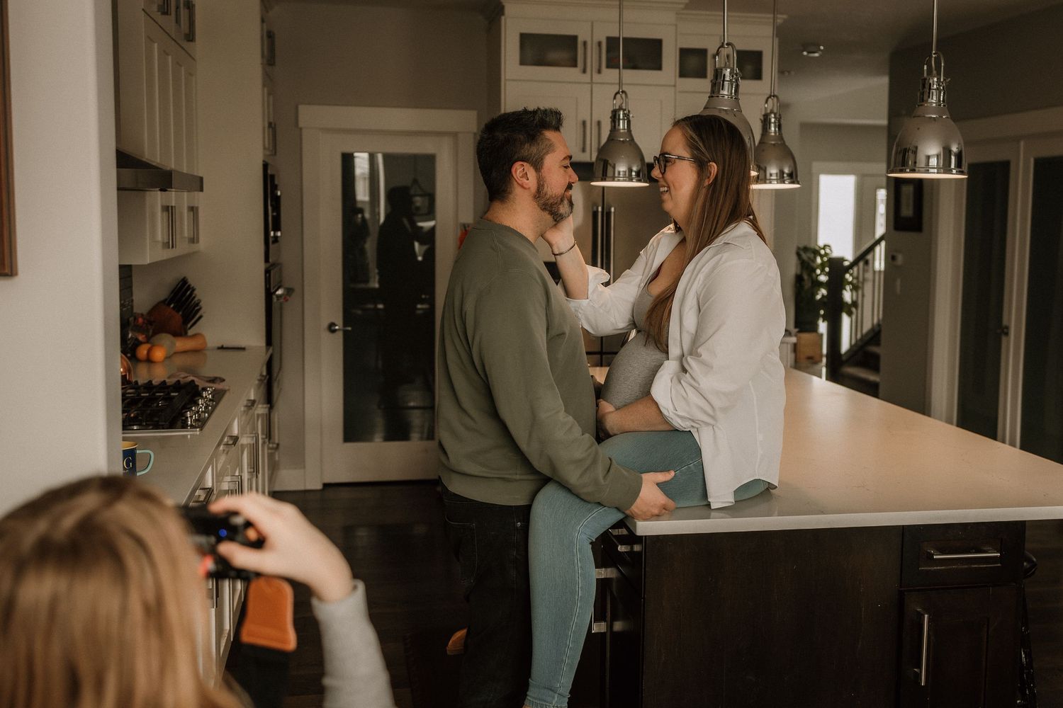 Couple sharing a tender moment in a kitchen while a photographer captures their maternity session.