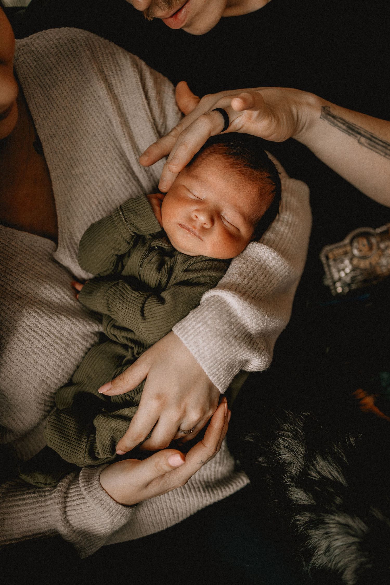 An in-home lifestyle session of a sleeping newborn baby in green outfit cradled in parents arms, with a gentle hand resting on its head.