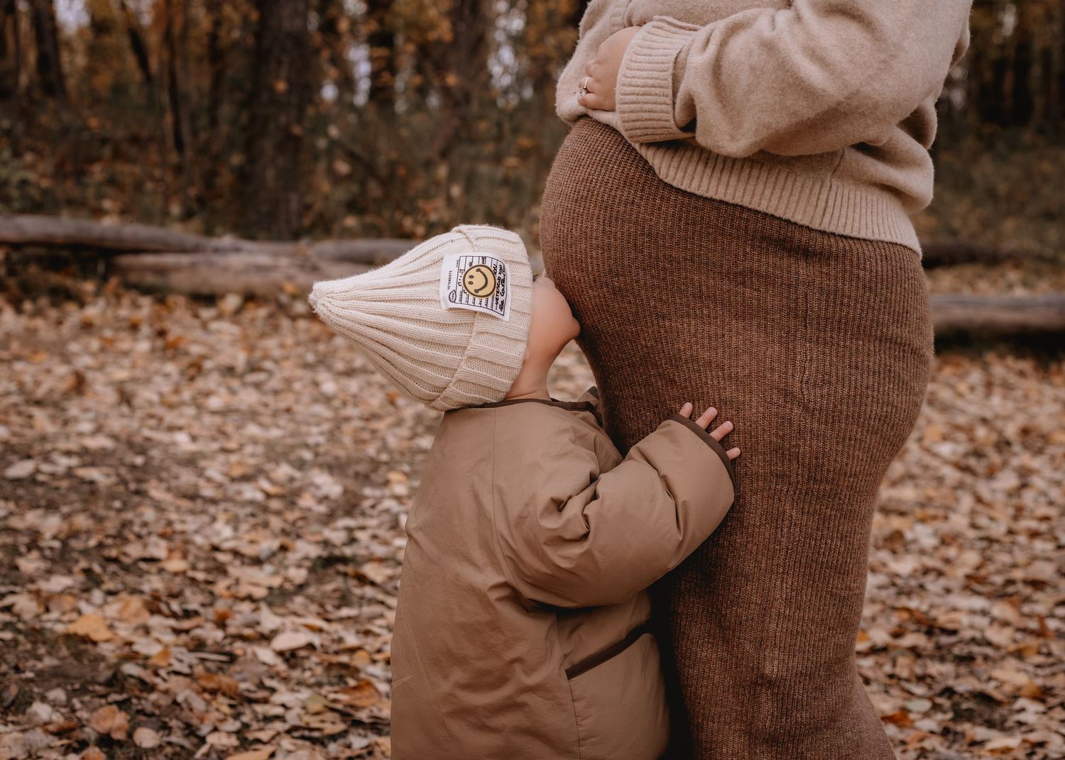 A toddler in a beige puffer jacket and knit hat hugs their pregnant mom in outdoor Calgary among autumn leaves.