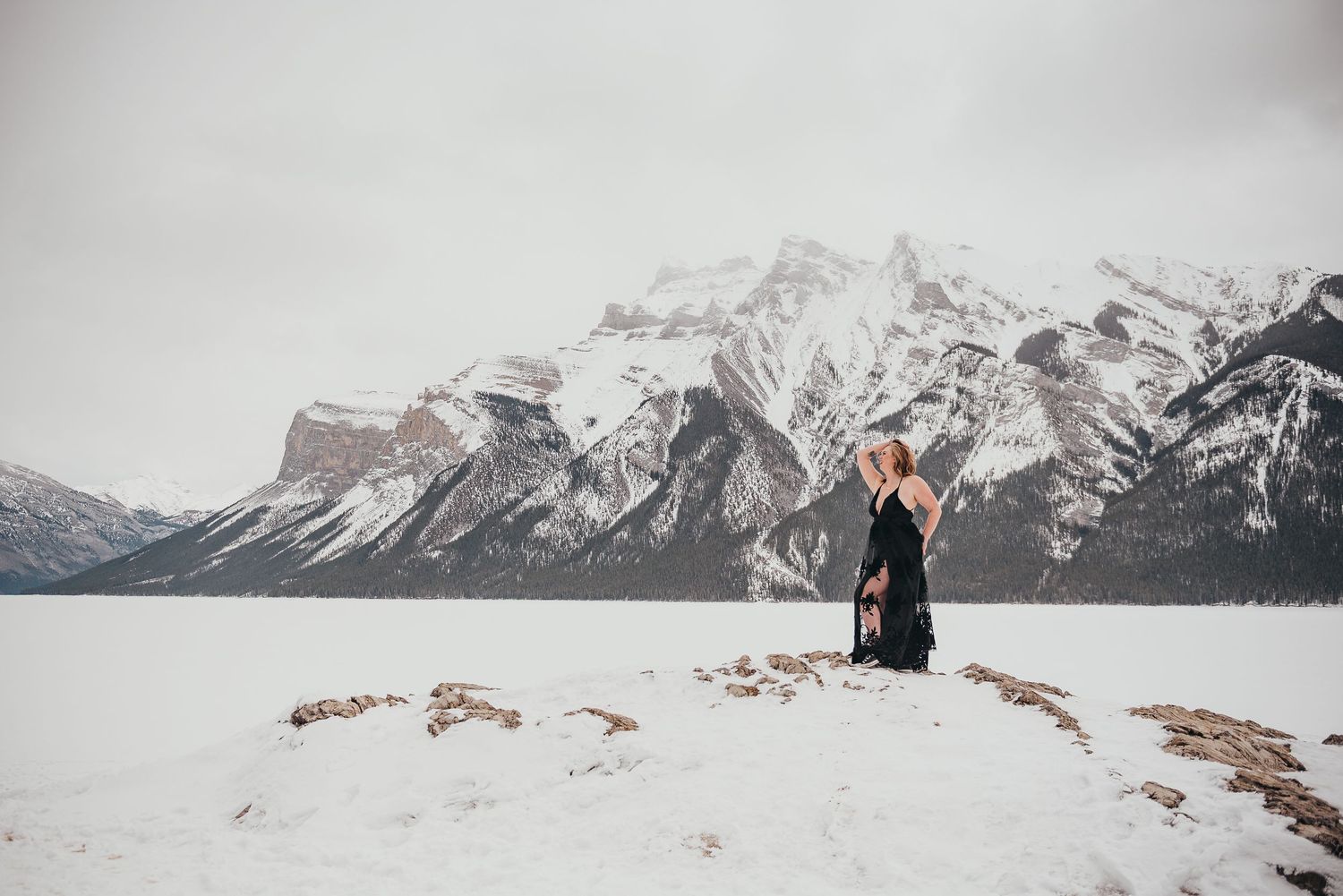 Woman in black dress poses on snowy shore with dramatic snow-capped Rocky Mountains in background.