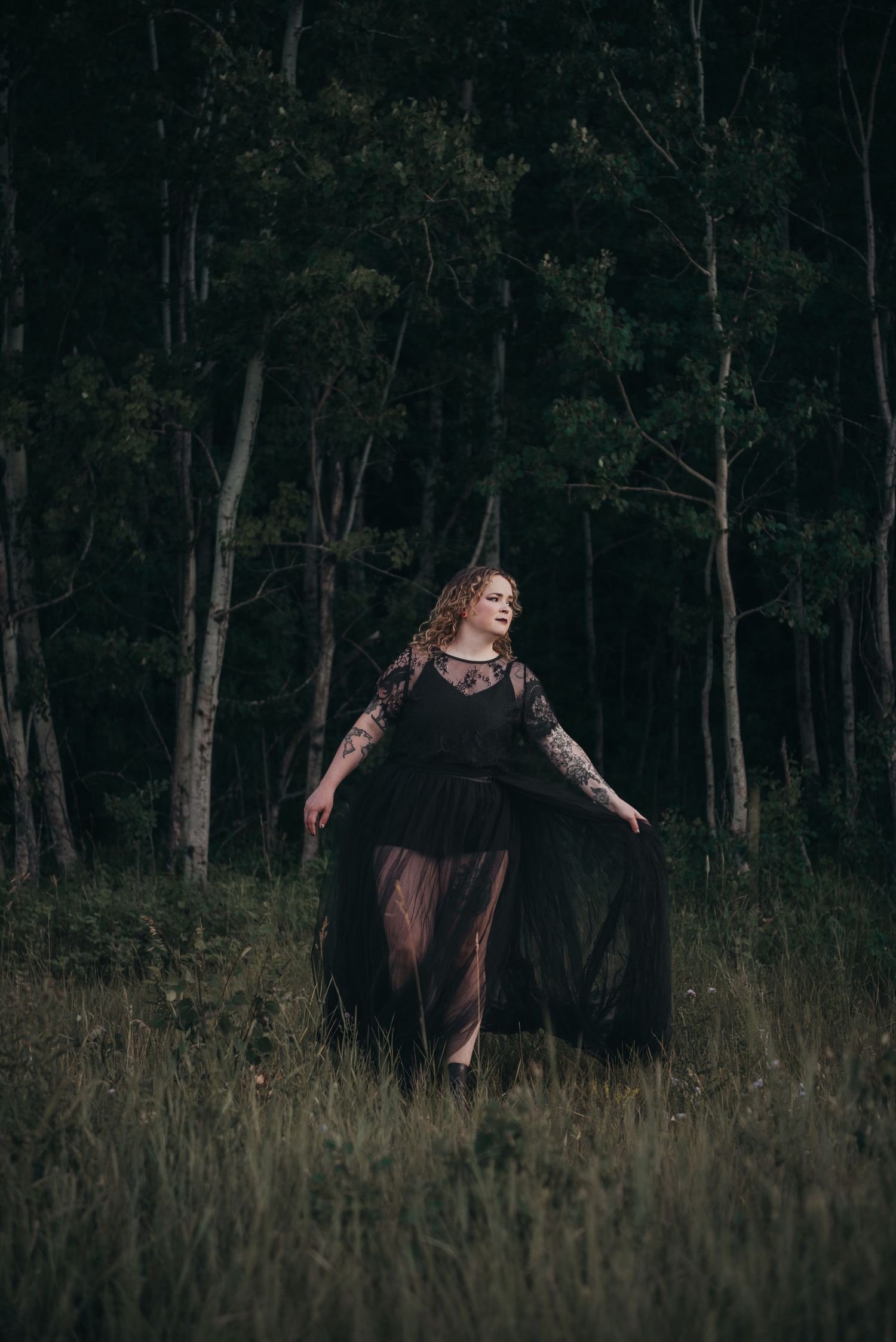 Woman in black lace gown standing in dark forest clearing at dusk, ethereal gothic fashion photography.