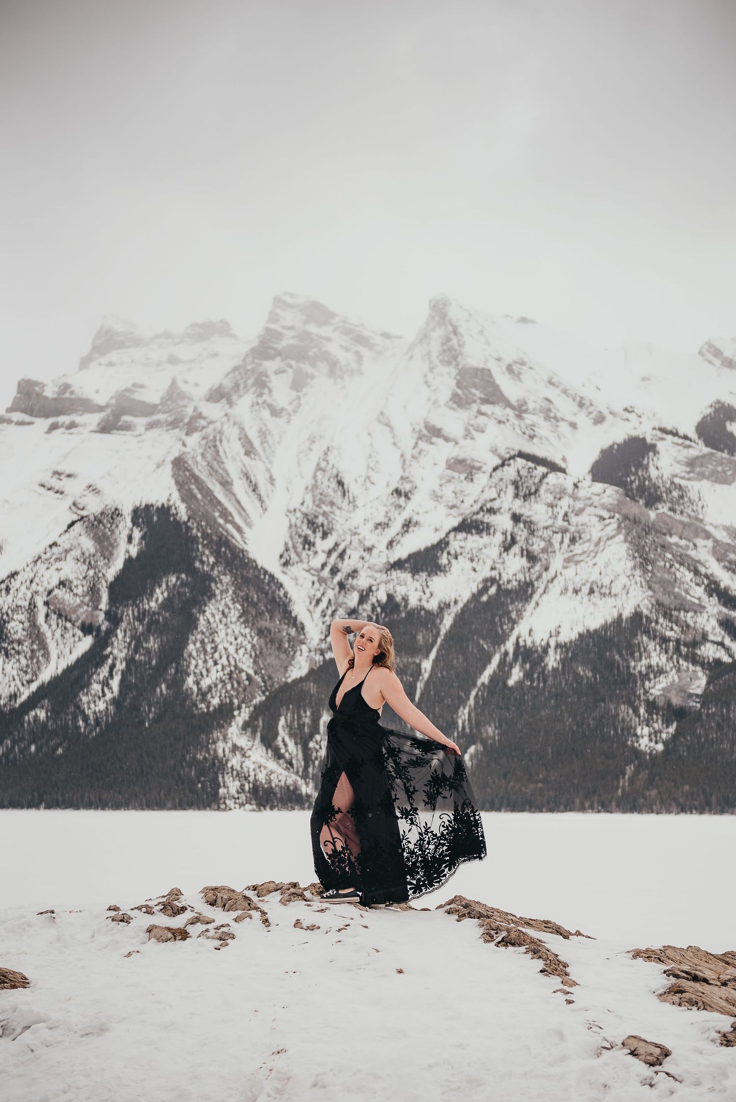 Woman in black lace dress poses gracefully on snowy landscape with dramatic Rocky Mountains in background.