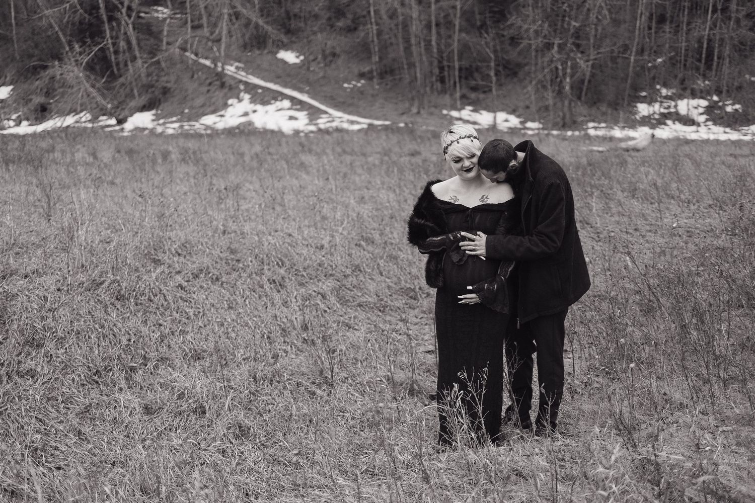 Black and white maternity photo of couple embracing in a field, woman in elegant black dress holding baby bump.