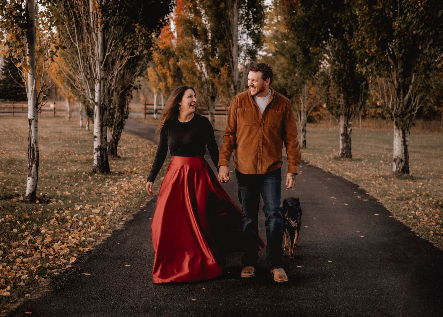 A couple strolls down a leaf-covered path surrounded by autumn trees.
