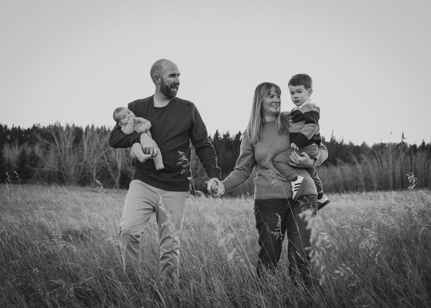 A family of four stands together in a grassy Calgary, AB field with trees in the background at dusk, captured in black and white.