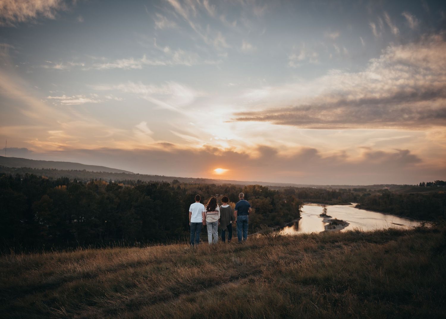Four people stand on a hill overlooking a river in Calgary and forest at sunset under a dramatic cloudy sky - family photographer