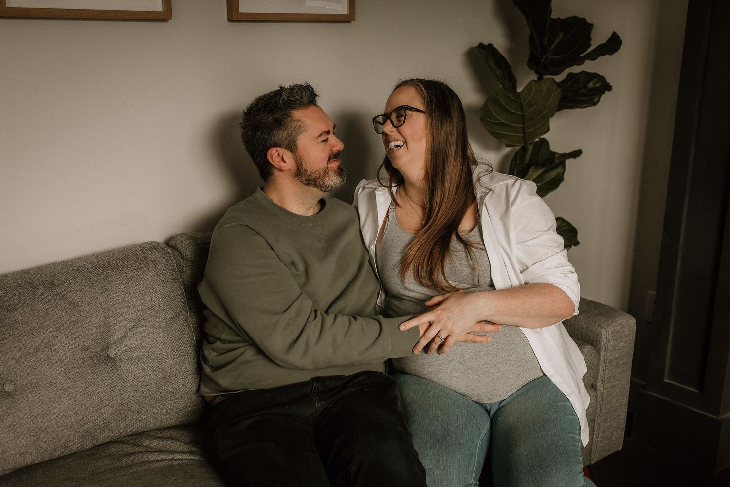Expecting couple sitting on a gray sofa, smiling at each other during an in-home maternity photo session.