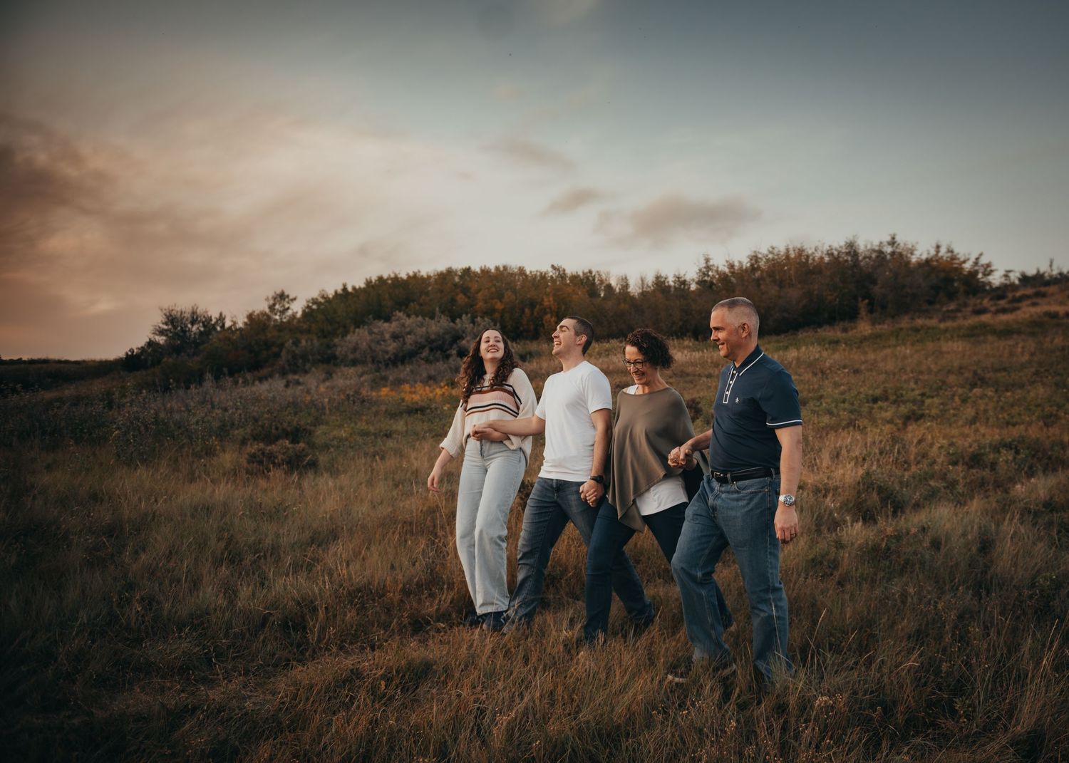 Family of four walks through golden meadow at sunset with dramatic cloudy sky backdrop during family photography session