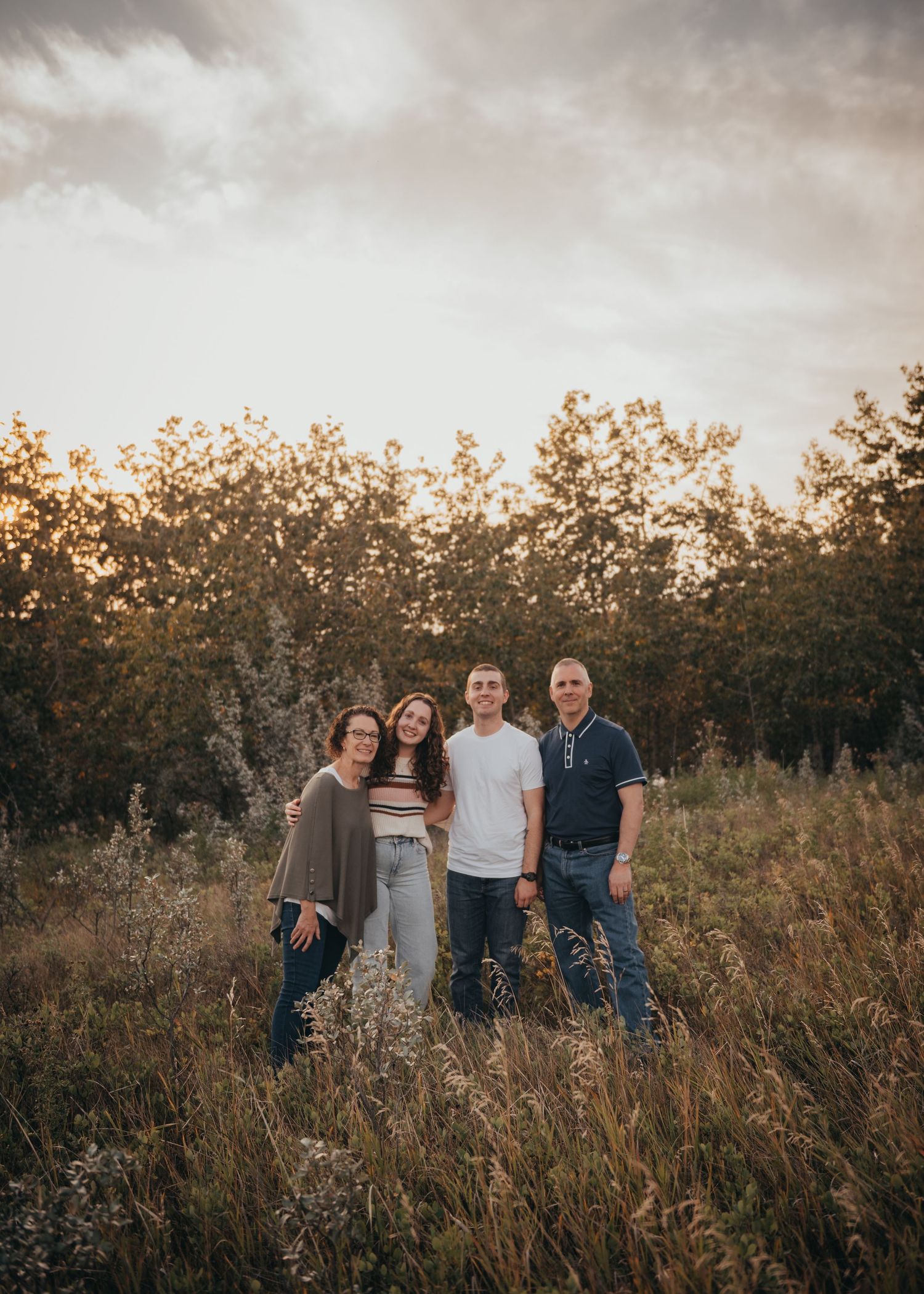 Family of four smiling together in a golden Calgary field at sunset, surrounded by wildflowers and trees.