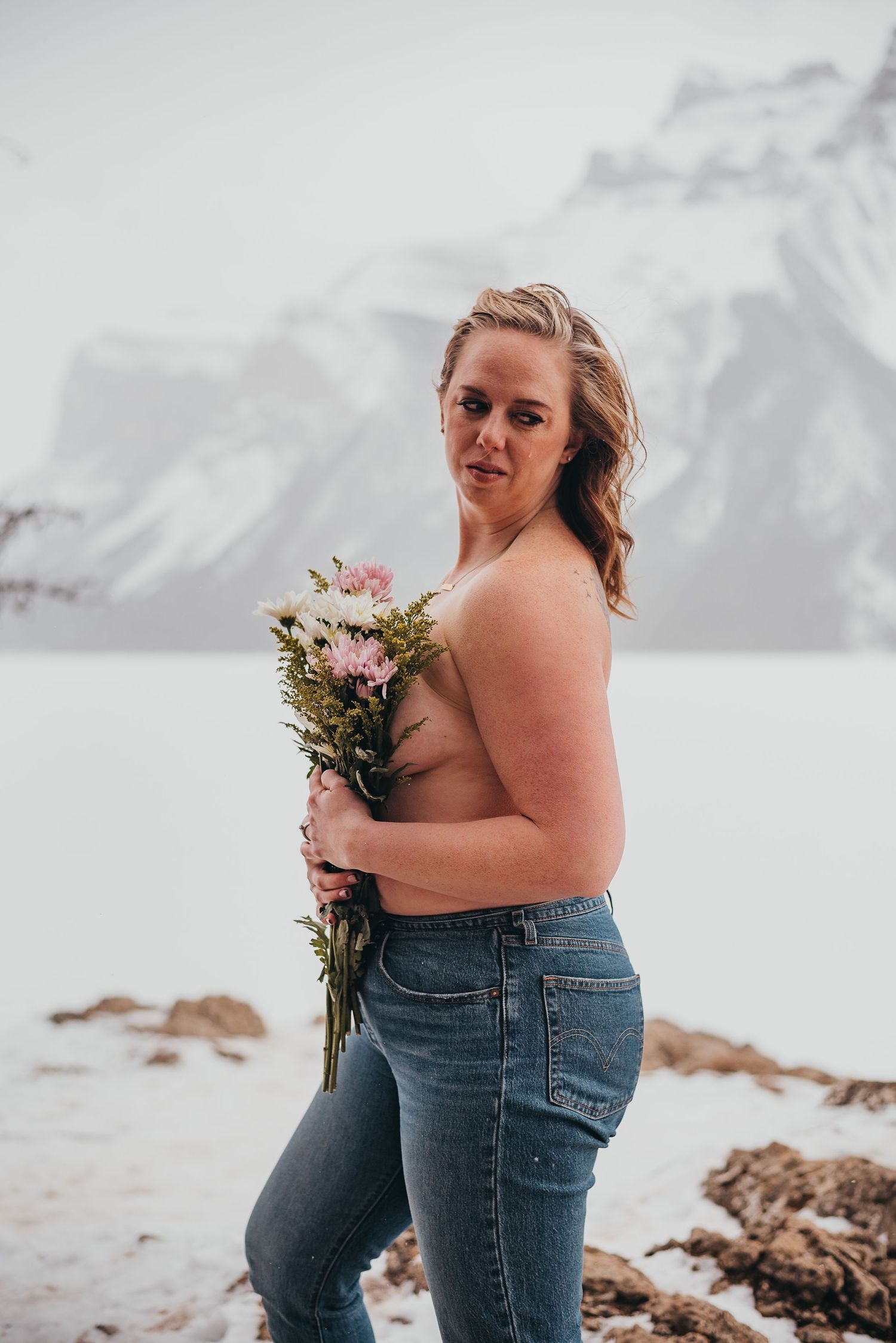 Woman holding pink flowers bouquet in jeans poses against snowy mountain landscape backdrop.