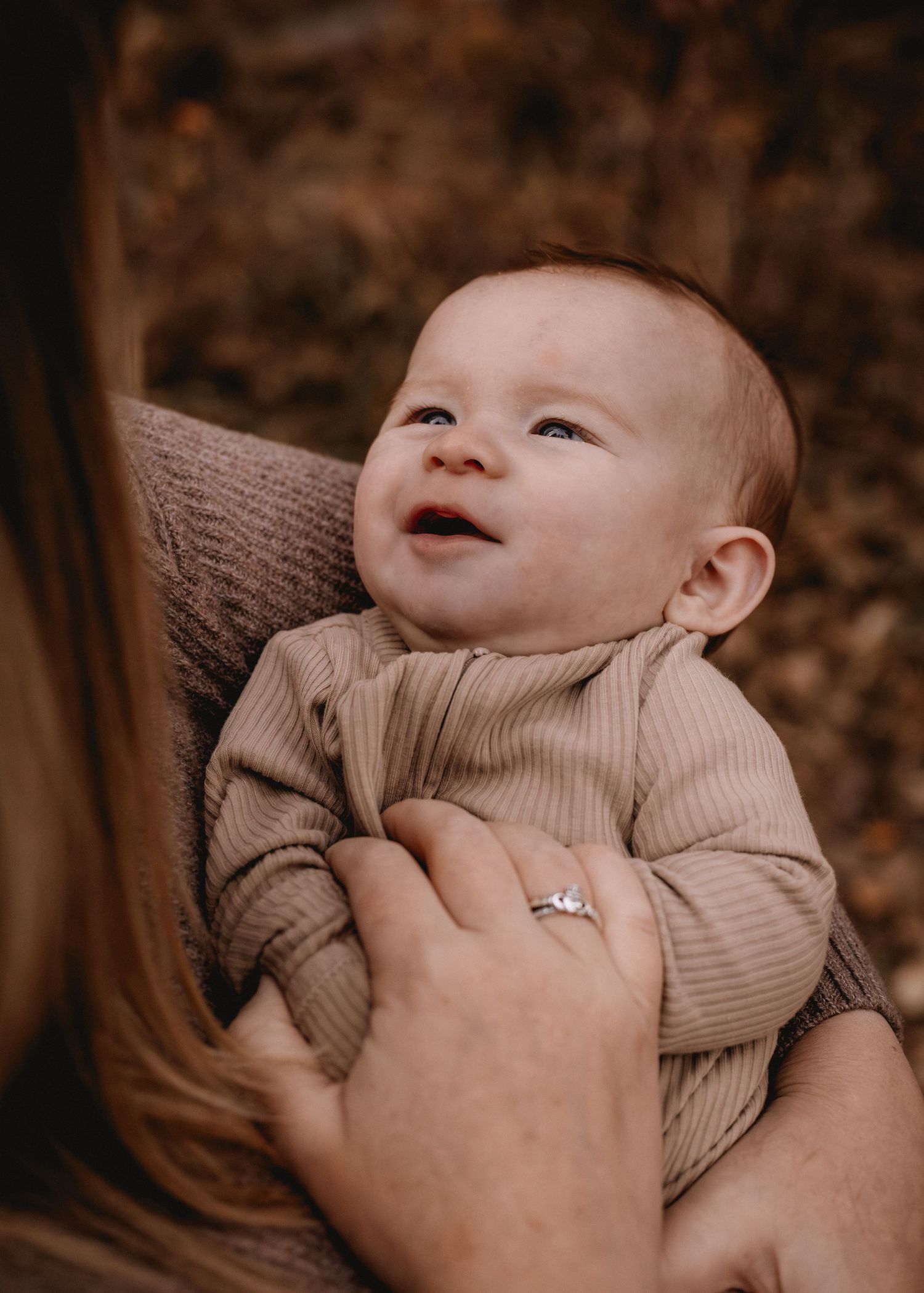 A smiling baby in a ribbed tan onesie being held by a mother wearing a diamond ring outdoors.