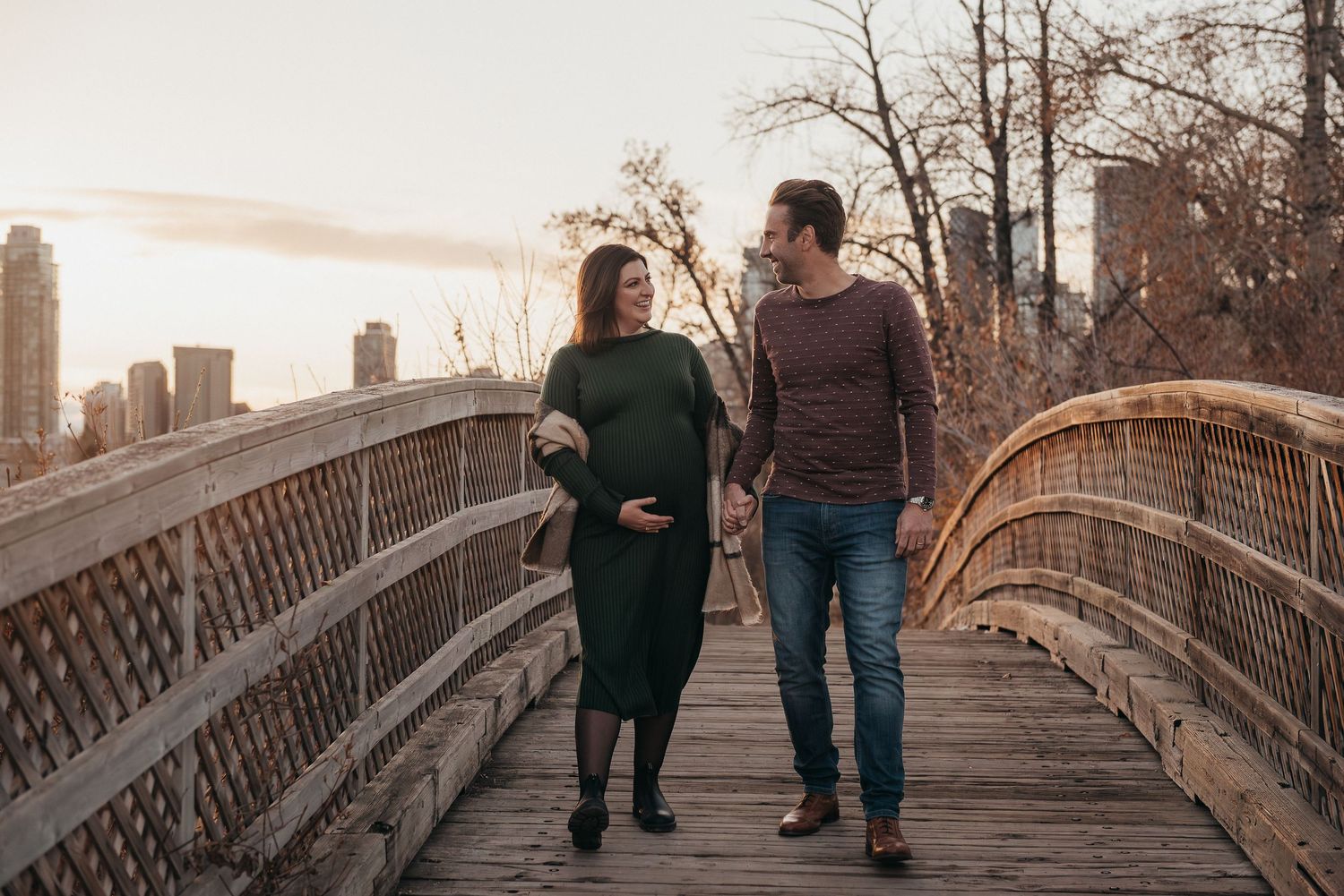 Pregnant couple walking hand-in-hand on a wooden bridge in Calgary, AB, with city skyline at sunset.