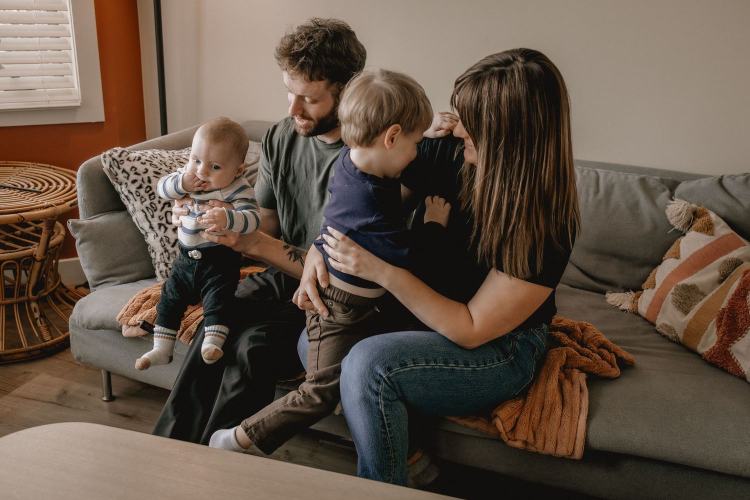 A young family with two small children sits together on a gray couch in a cozy living room, sharing a warm moment for Calgary photographer