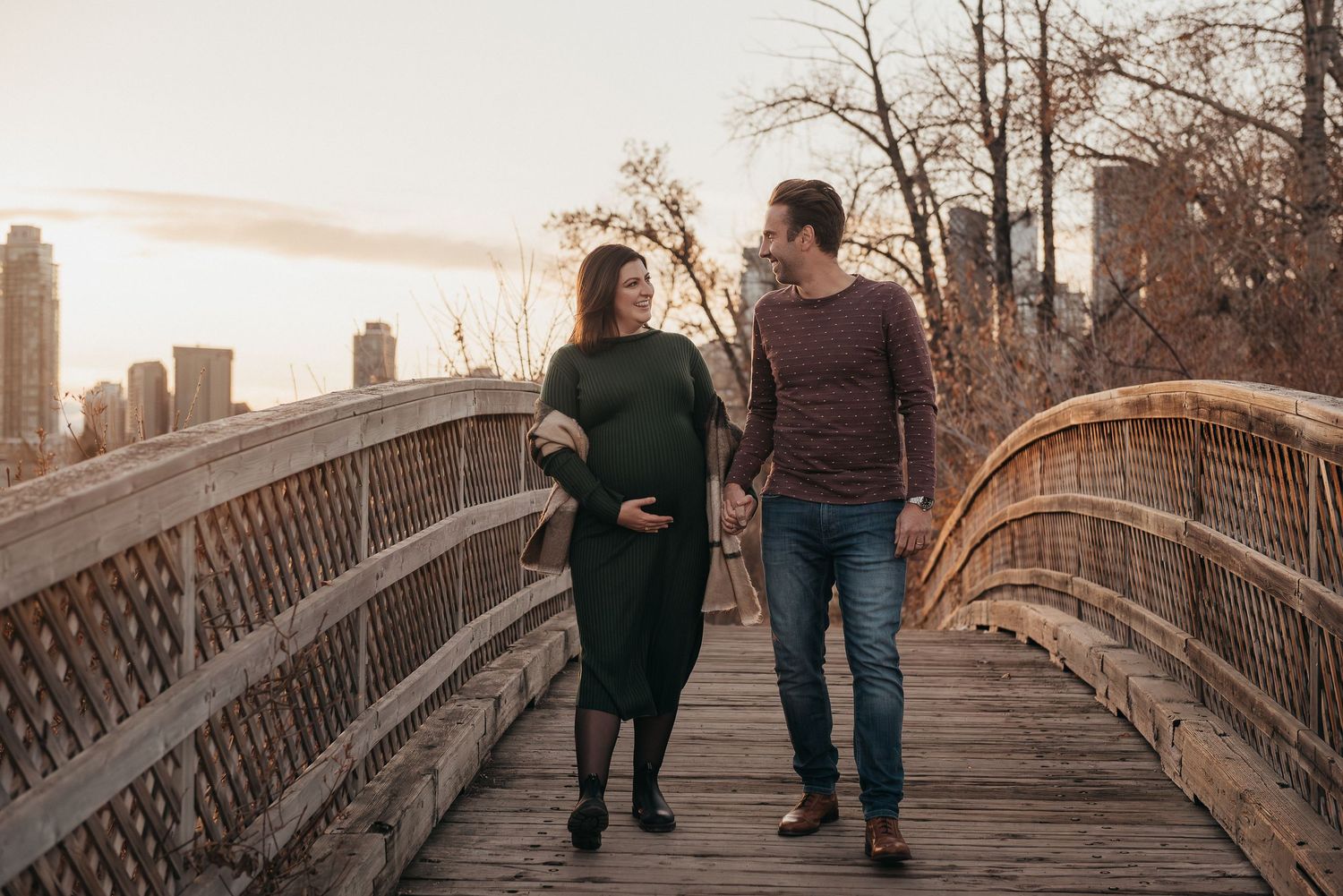 Maternity session couple walking hand in hand across wooden bridge with Calgary's city skyline in background.