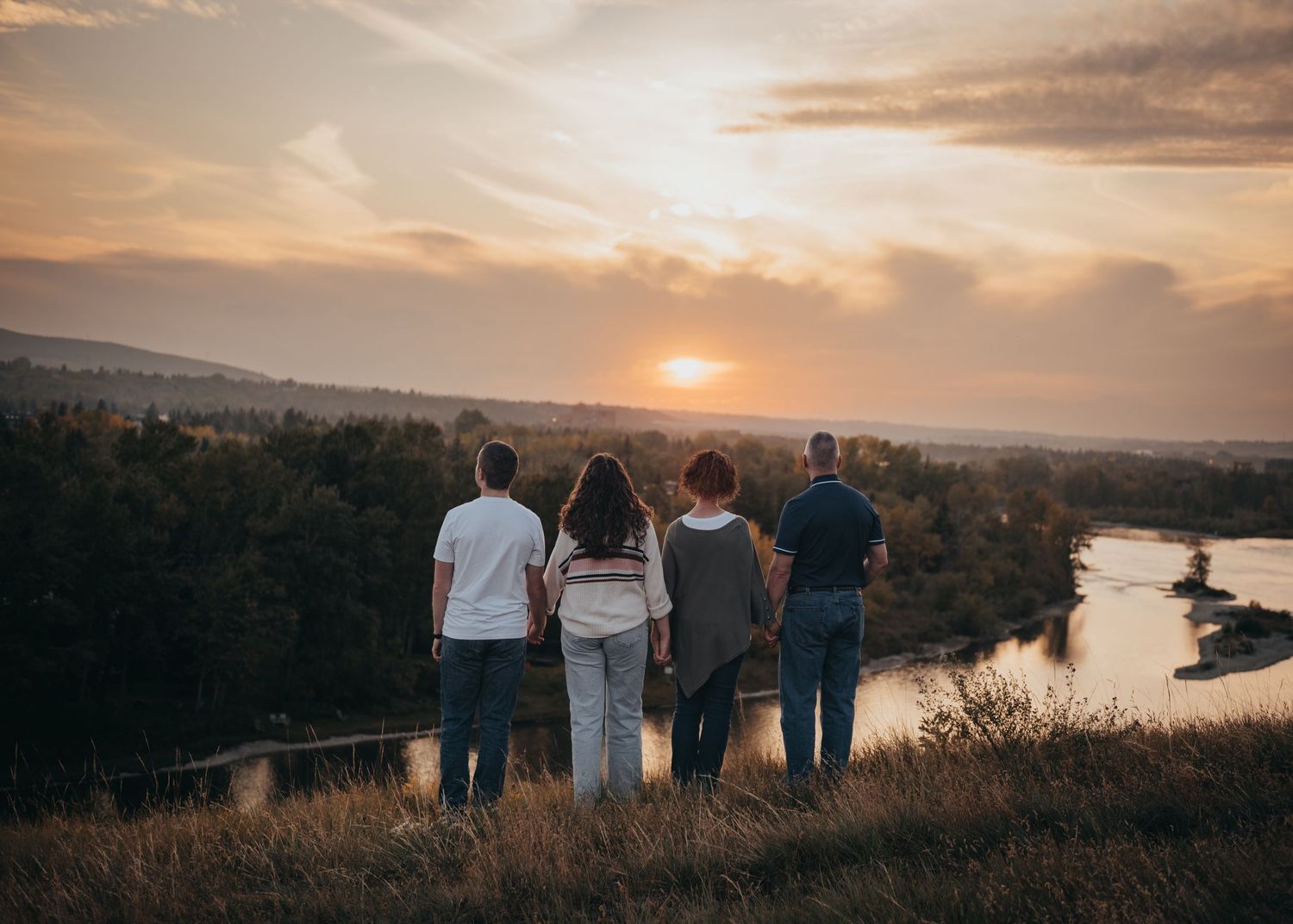 Family of four stand together on a hilltop overlooking a 
Calgary river valley at golden sunset.