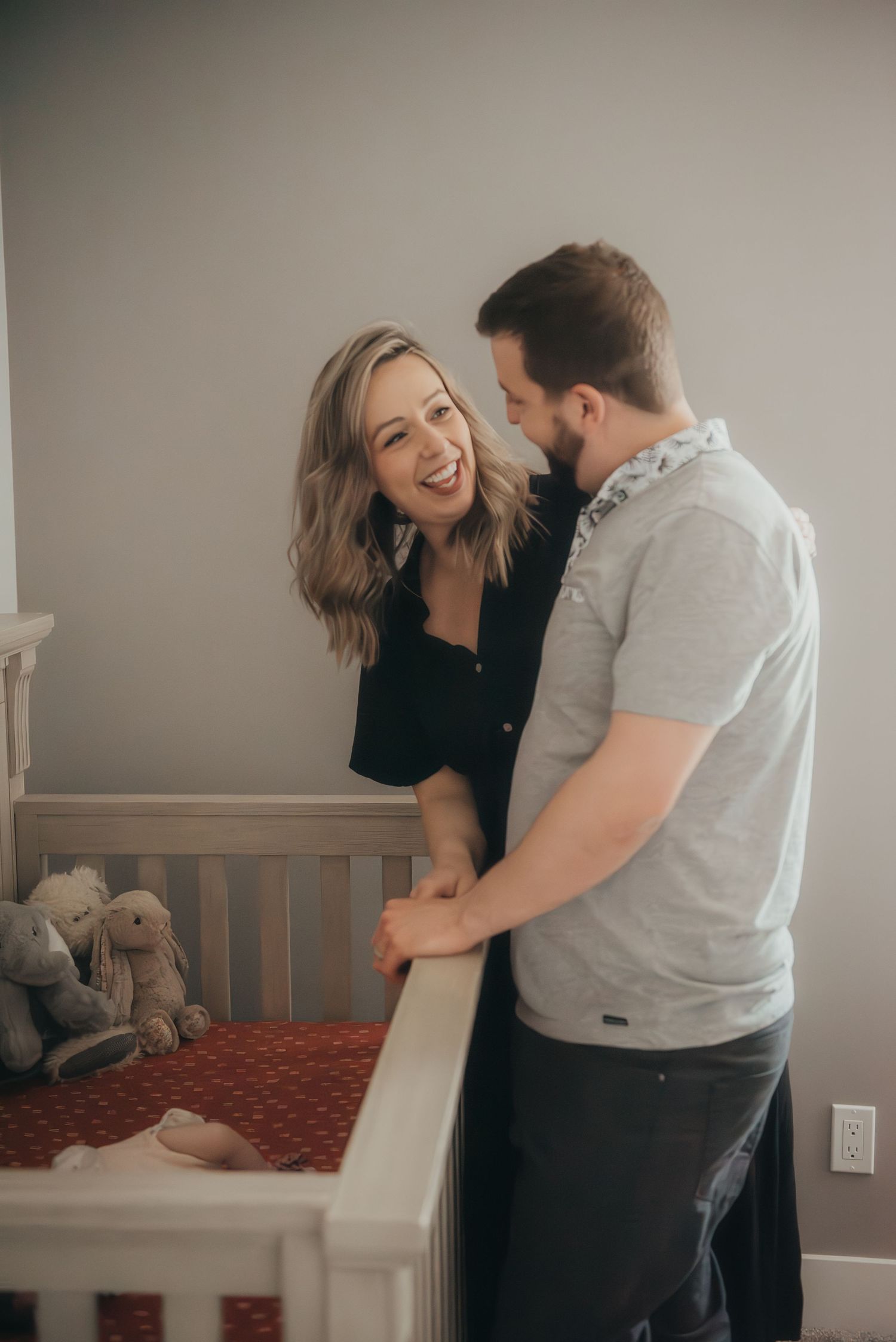 A smiling couple stands together over a white baby crib sharing a joyful moment in a nursery with their newborn