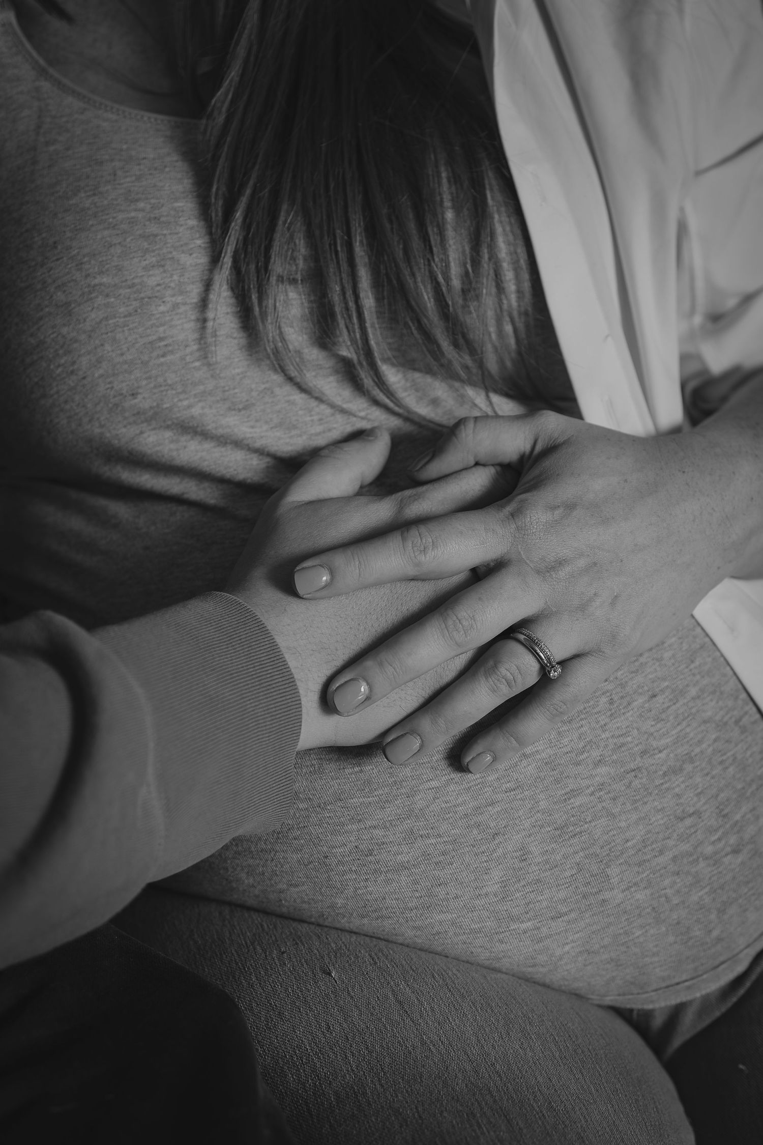 In-home maternity session black and white photo of a couple holding hands on a pregnant woman's baby bump, with a wedding ring visible.