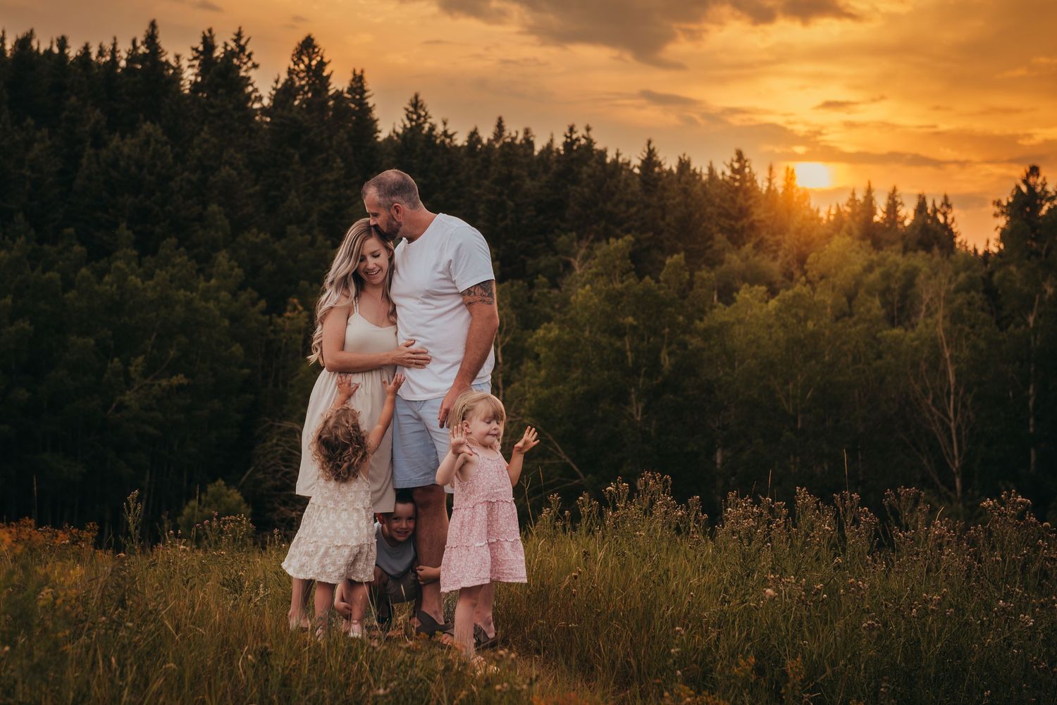 Family of five posing at golden sunset in a Calgary meadow with forest backdrop, parents and two young daughters in white and pink dresses.