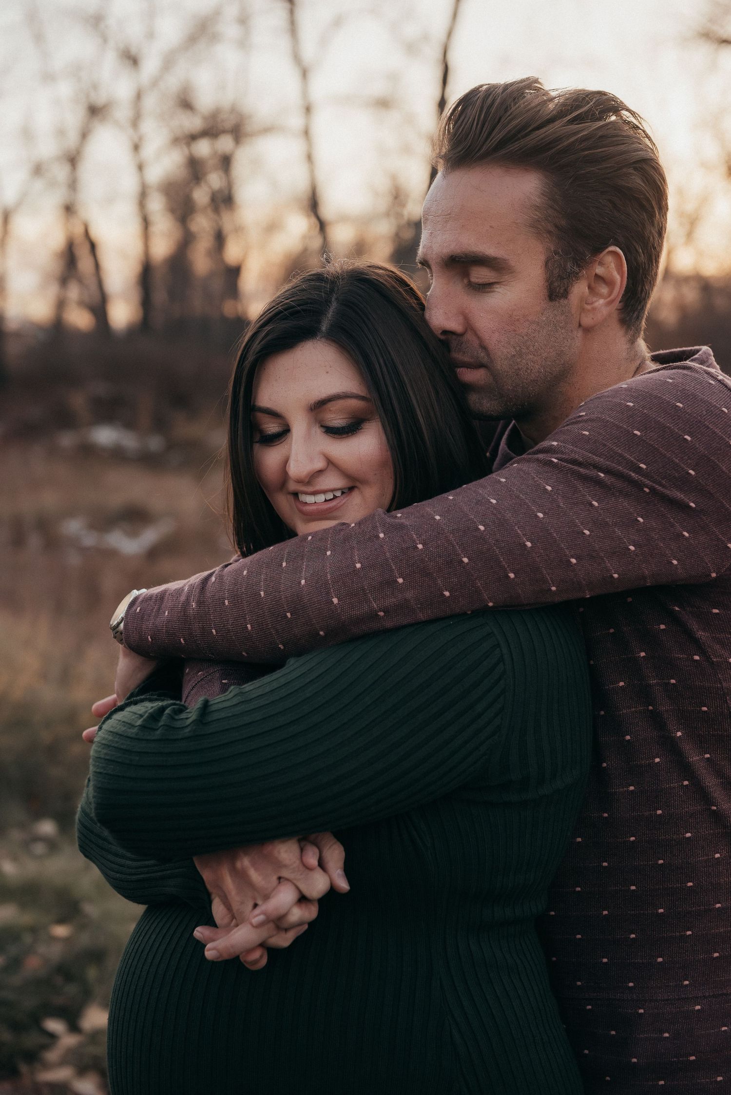 A couple shares a warm embrace outdoors at sunset, man hugging woman from behind, both smiling peacefully during maternity photoshoot