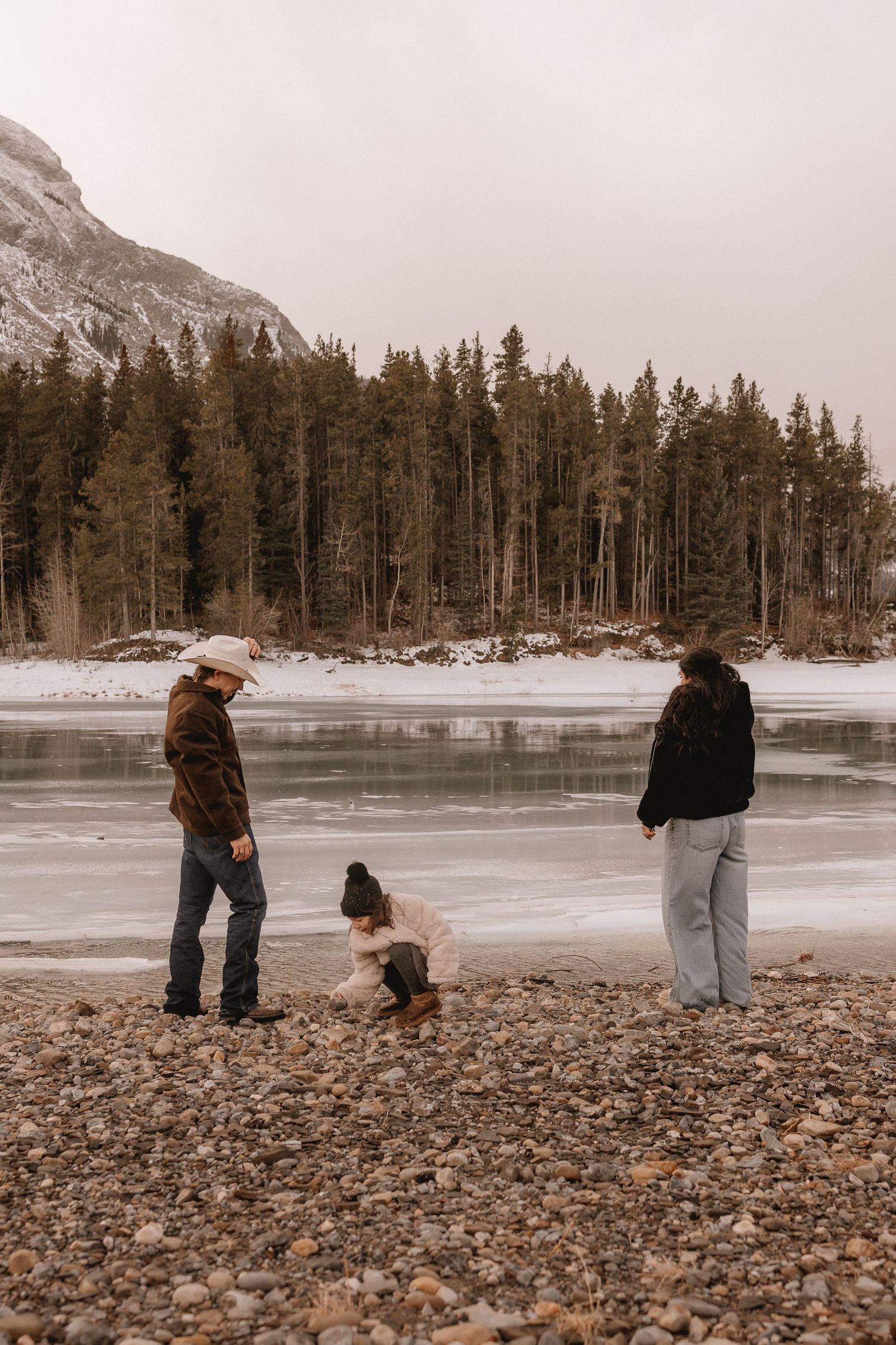 Calgary family of three stand at the edge of a frozen lake surrounded by evergreen trees and mountains in winter.