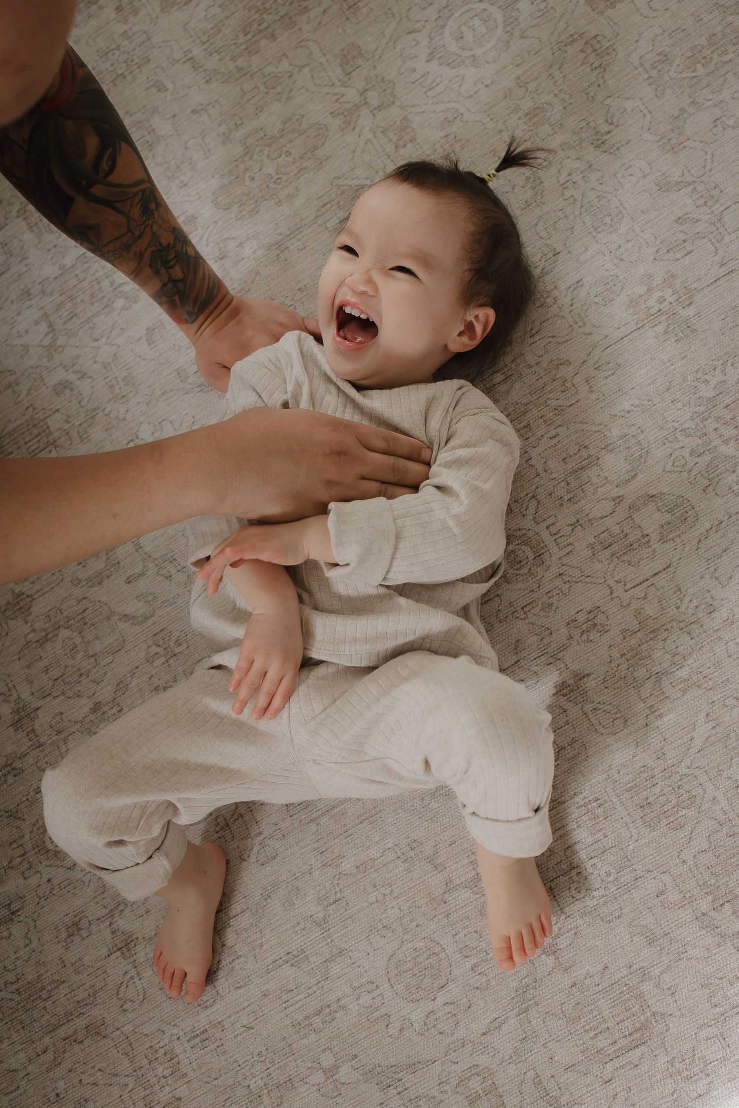 A laughing baby in a beige outfit lies on a patterned rug while her dad tickles them for a Calgary lifestyle family photographer.