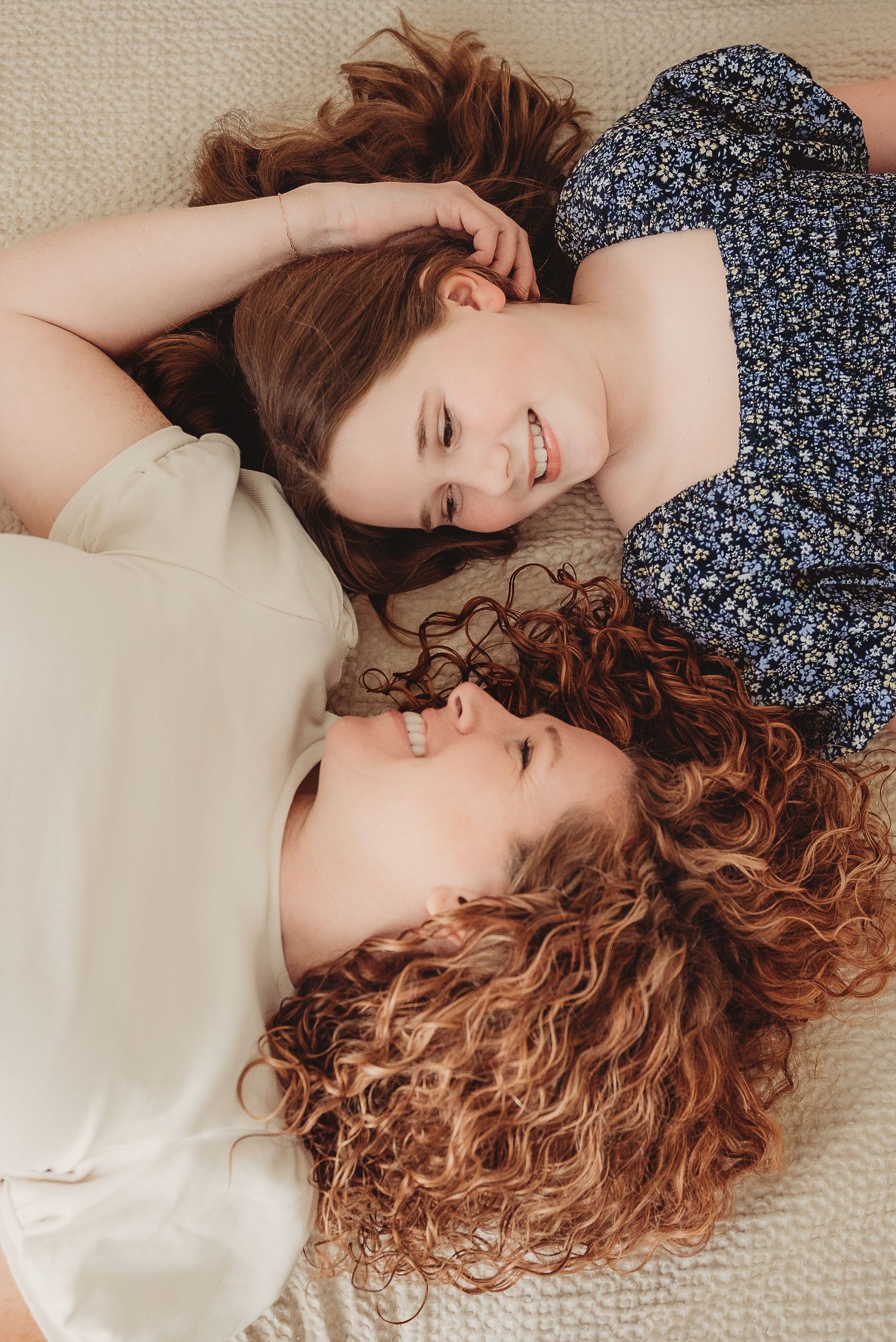 Mom and daughter lying on a light surface, heads together, wearing casual floral and cream outfits.