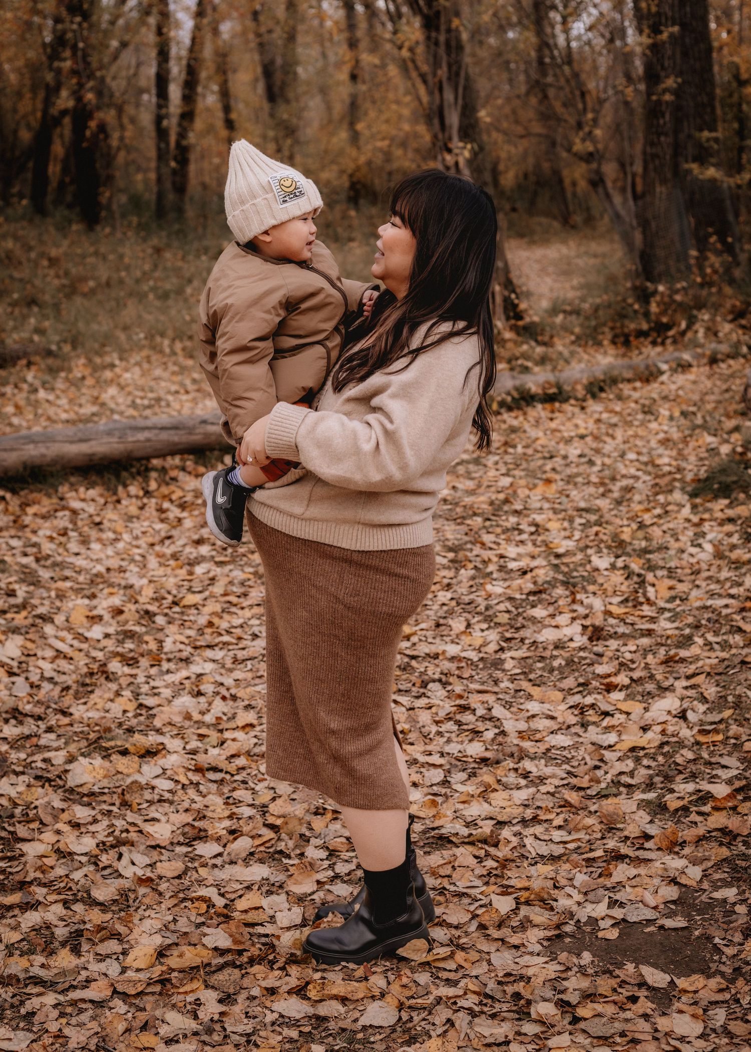 Woman in beige sweater and brown skirt holds toddler in white beanie among autumn fallen leaves in forest.