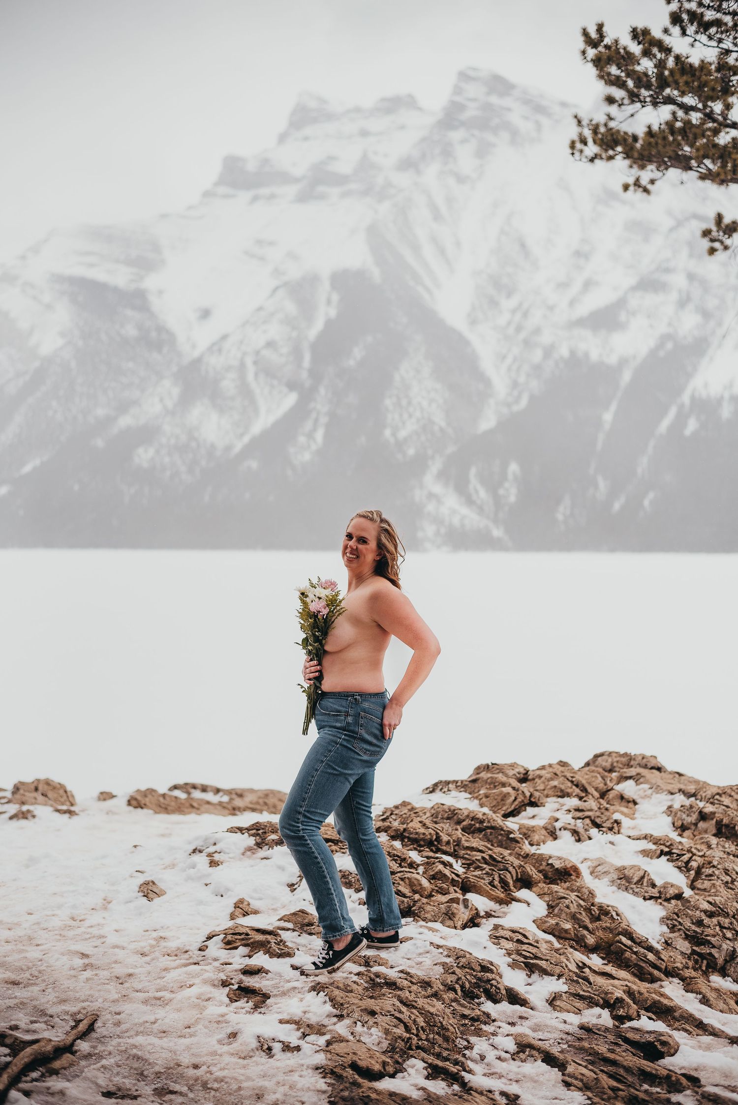 Topless woman holding flowers stands on snowy rocks with mountain Banff, AB lake backdrop in winter landscape.