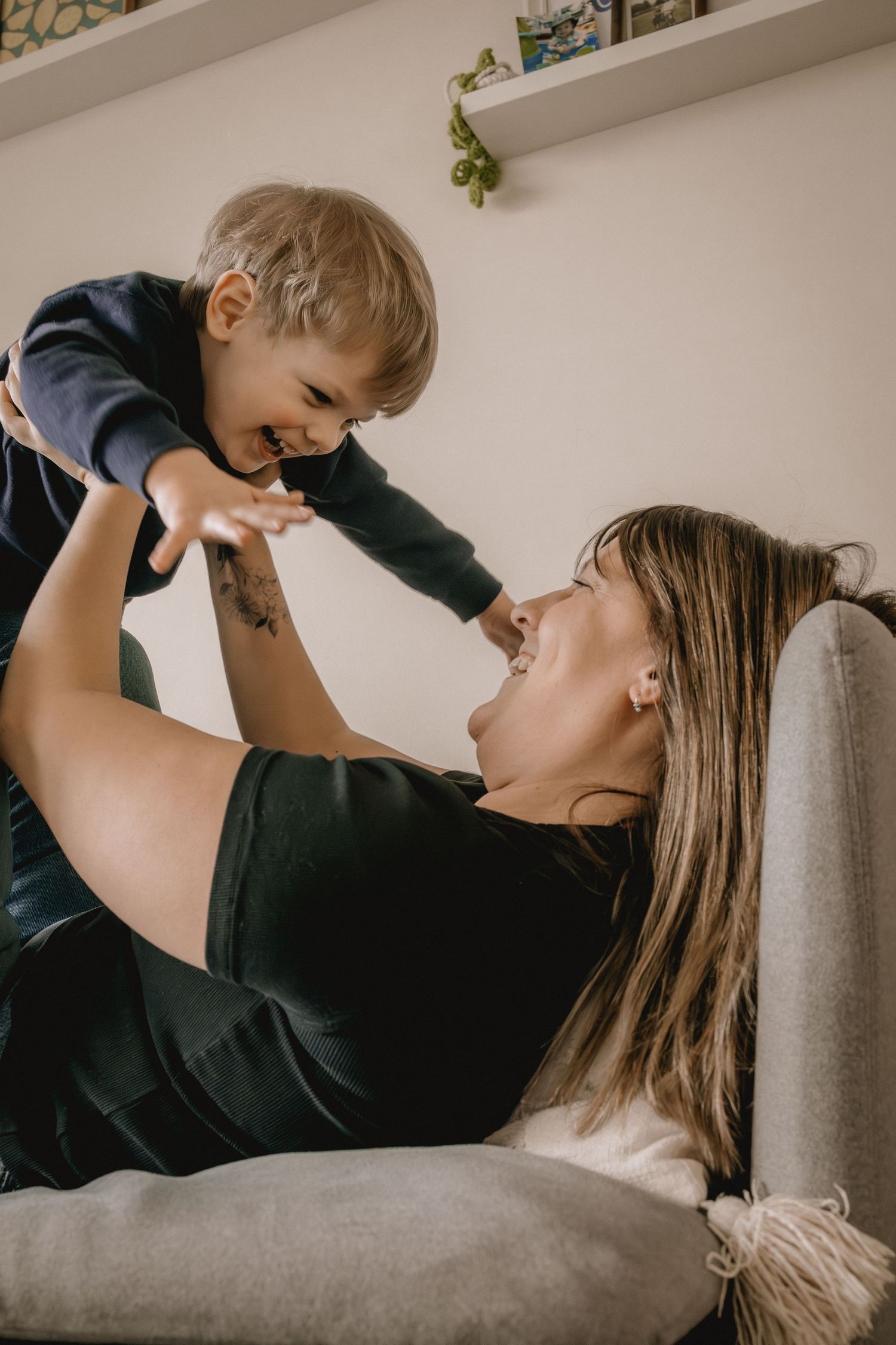 A Calgary mother joyfully lifts her smiling toddler above her while relaxing on a couch indoors for a family photographer