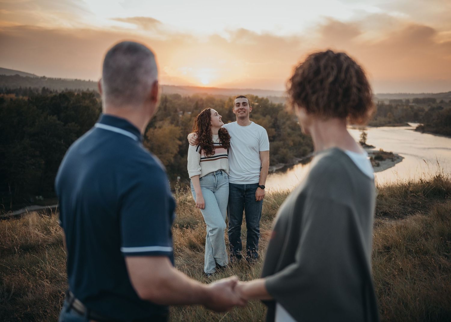 Mom and dad with their two children at sunset overlooking a scenic Calgary river valley with rolling hills in the background.